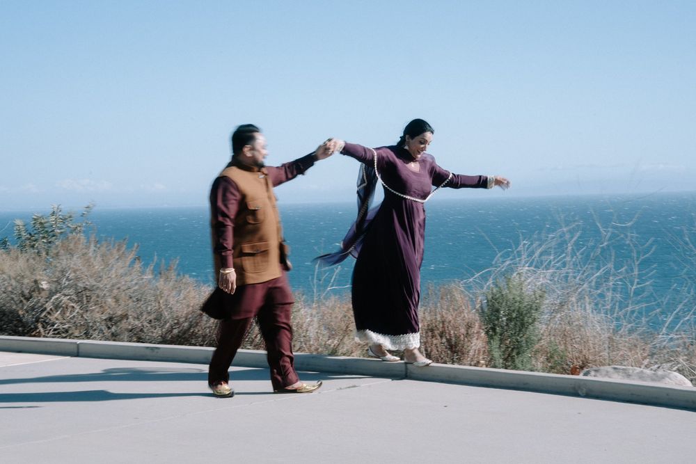 Two people hold hands while dancing together on a scenic coastal road overlooking the ocean on a sunny day.