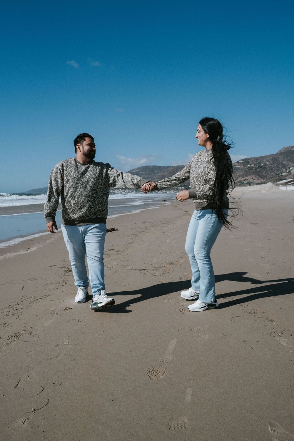 A couple in matching knit sweaters enjoying a casual stroll along a scenic beach with mountains in the background.