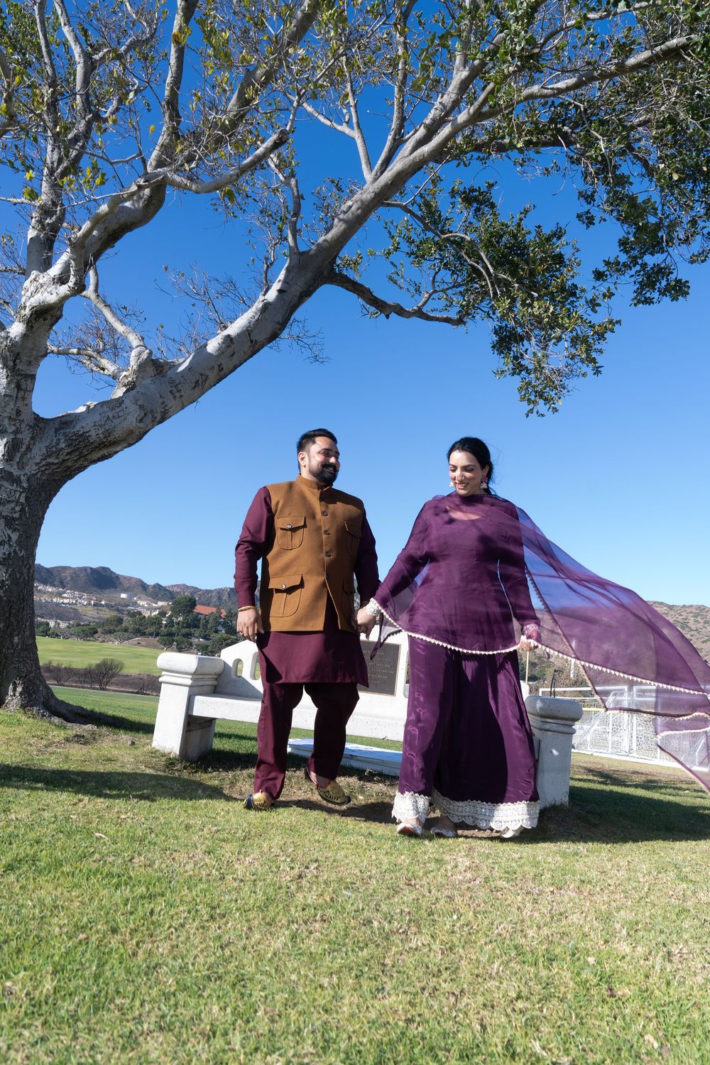 A couple in coordinated purple outfits posing under a large sycamore tree on a sunny day.
