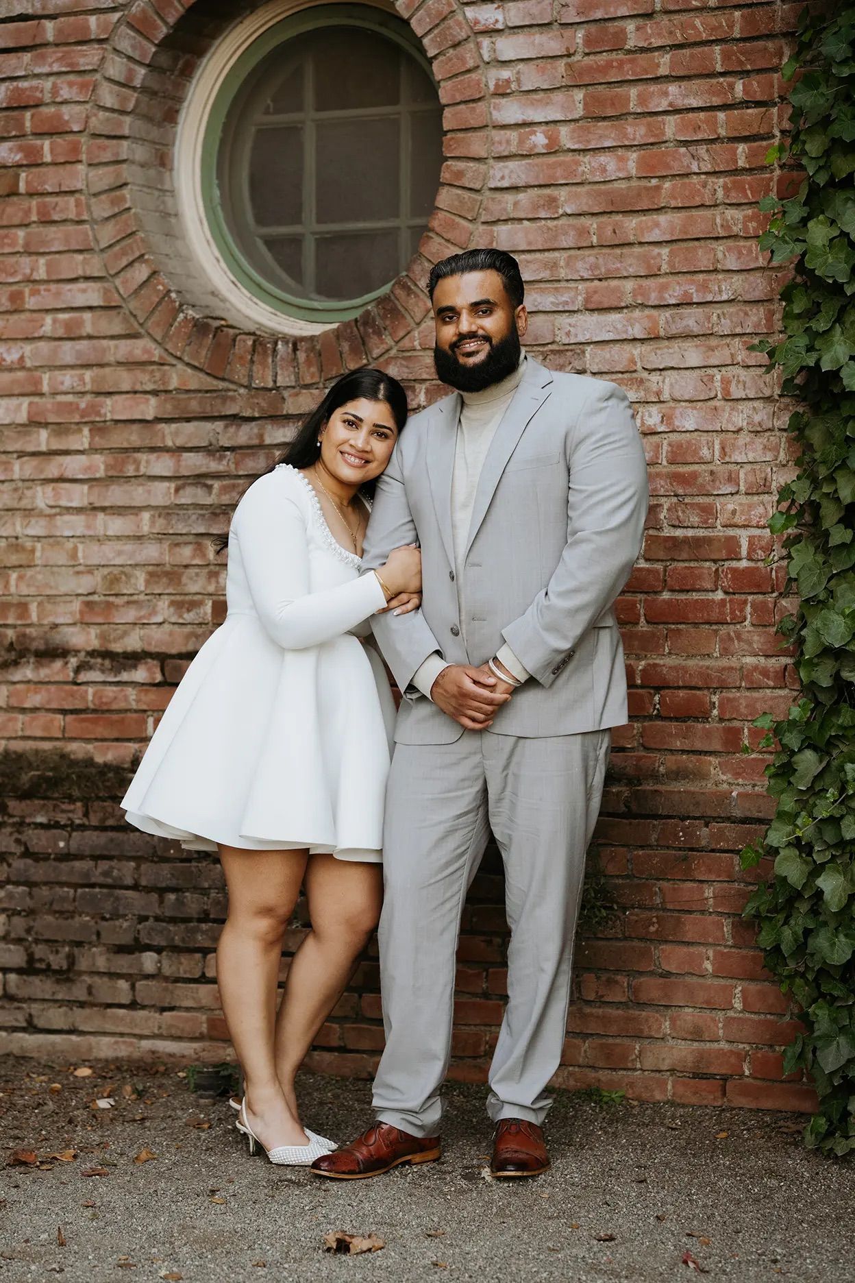 Couple posing against brick wall, woman in white dress and man in light gray suit, smiling outdoors.