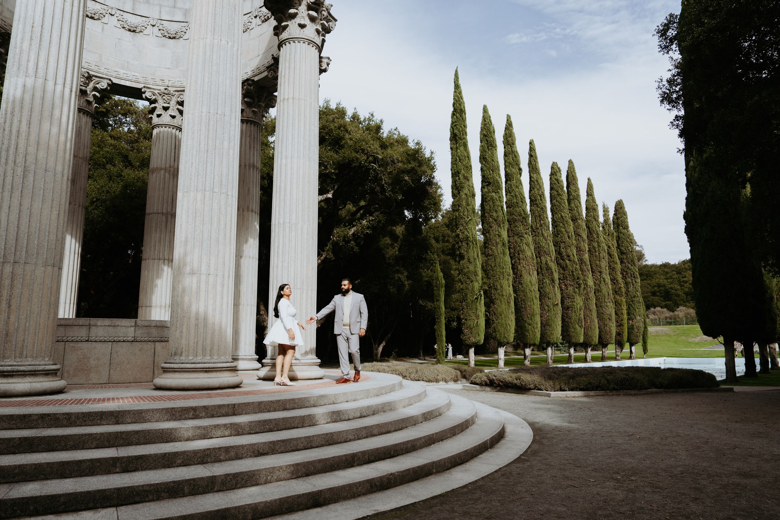 Couple holding hands on grand steps beside tall columns with cypress trees in background.