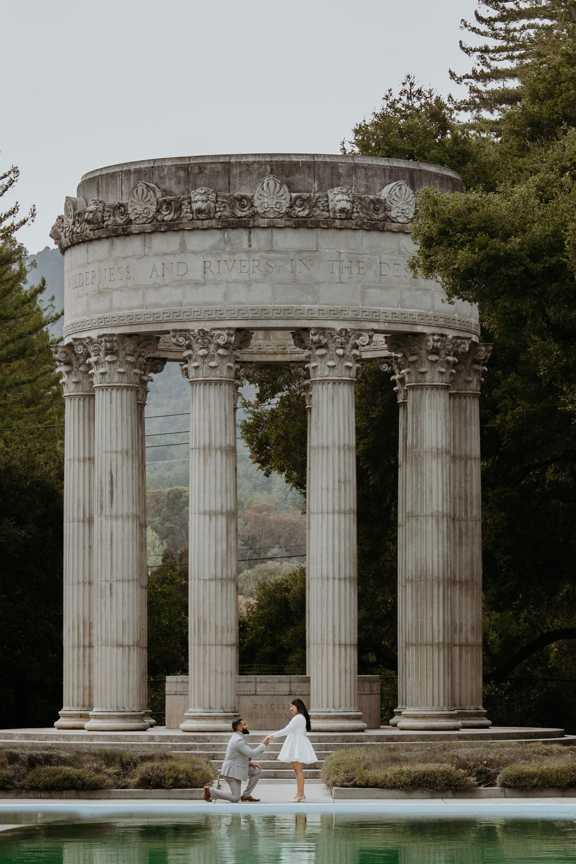 A couple shares a marriage proposal moment by a classical Roman-style colonnade beside a reflecting pool.