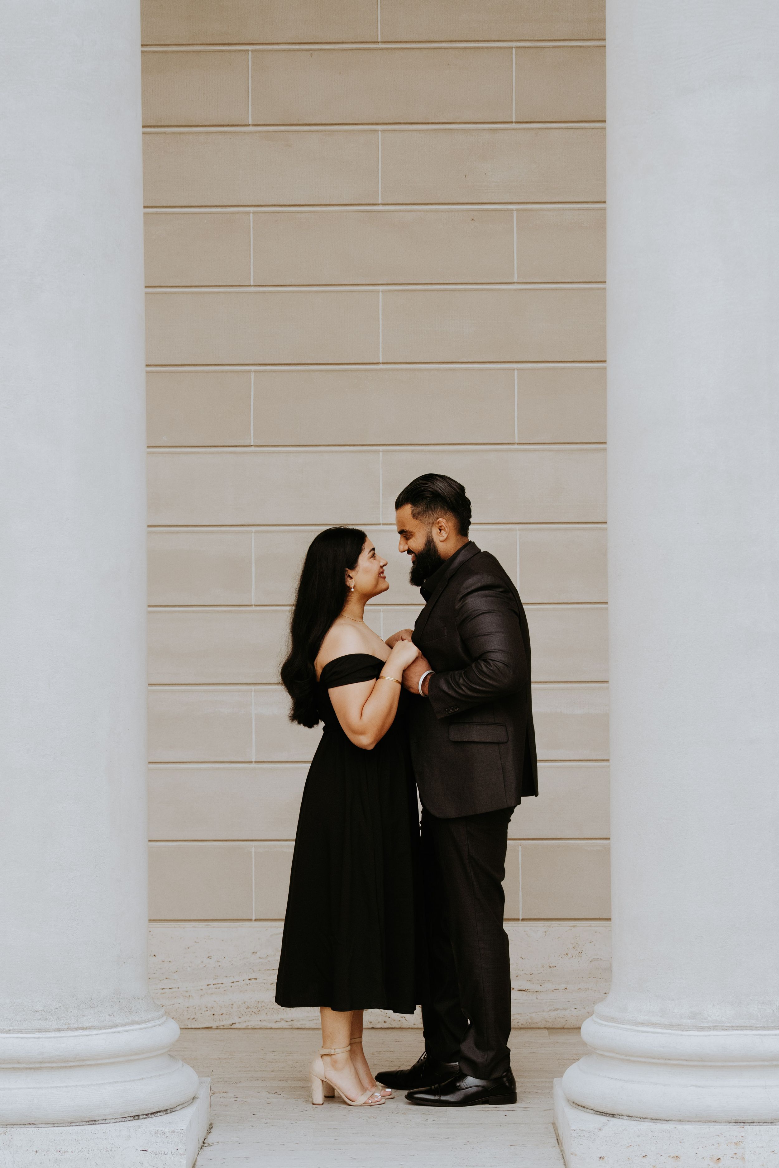 Romantic couple in black outfits embracing between elegant columns against a beige wall.