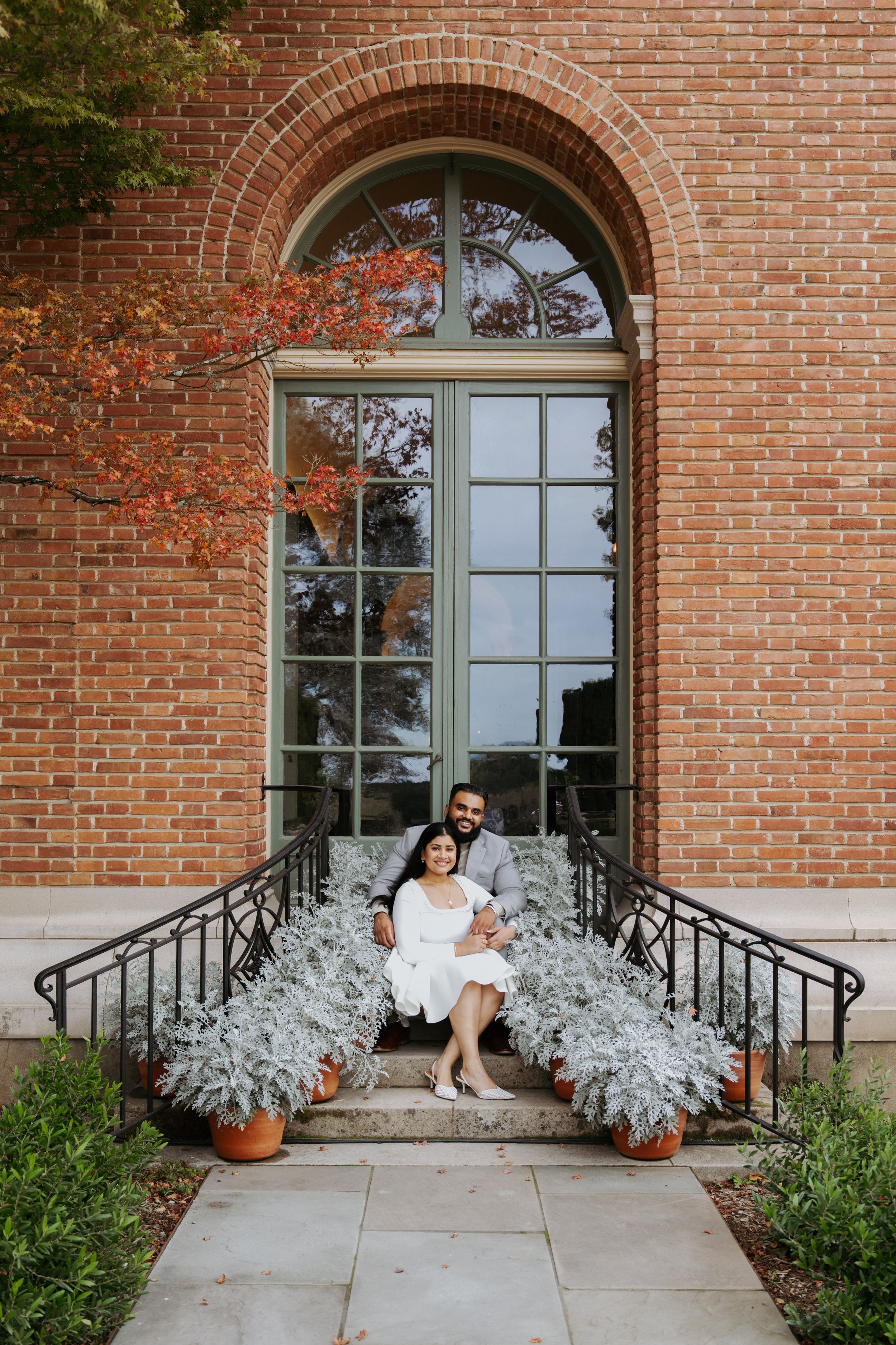 Couple sitting on brick building steps surrounded by silver foliage plants and autumn trees during engagement session.