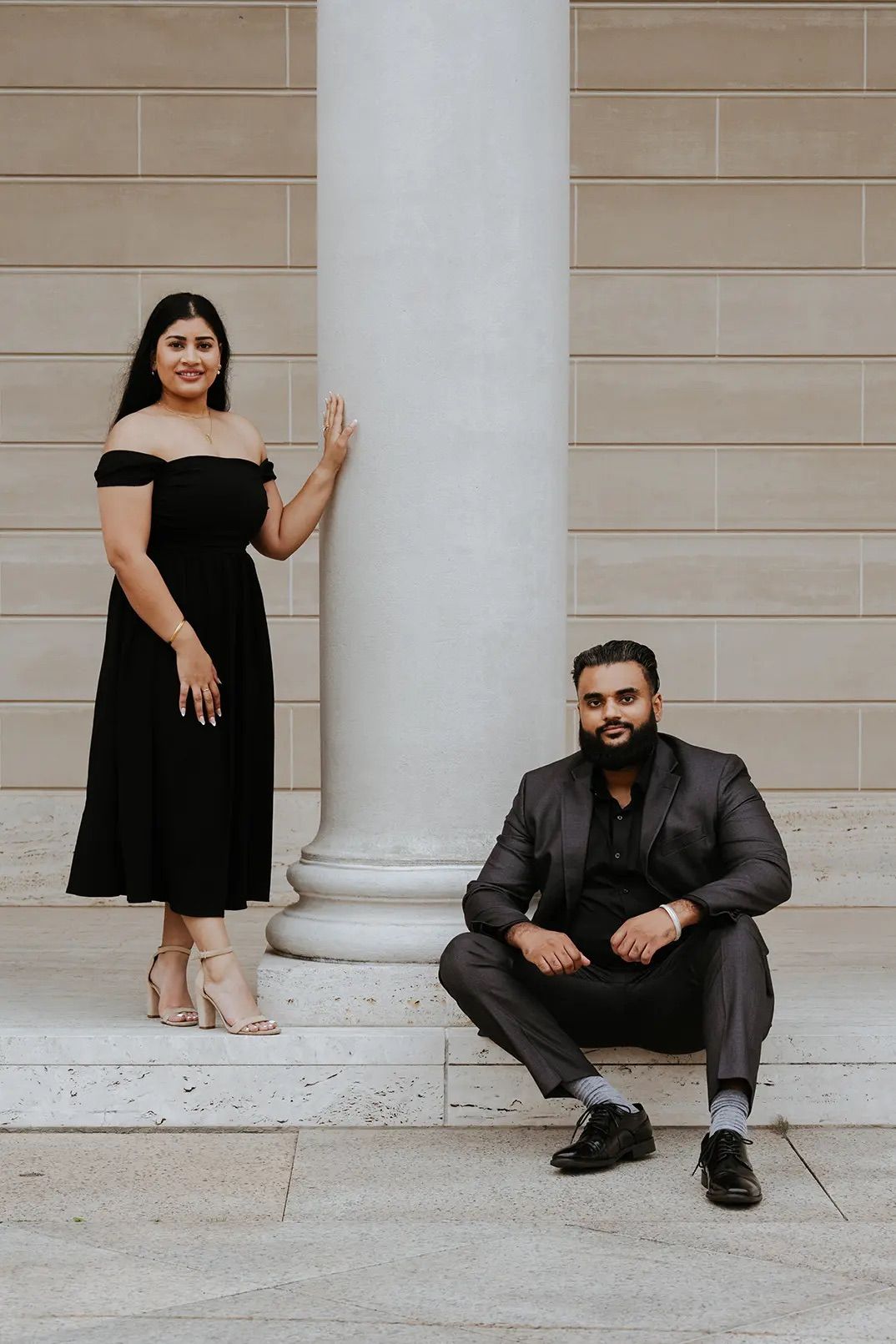Couple in matching black outfits posing by a white column on marble steps.