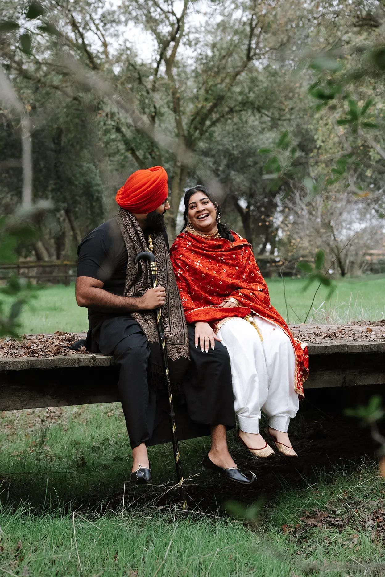 Sikh couple sitting on wooden bench in park, man in orange turban, woman in red dupatta smiling.