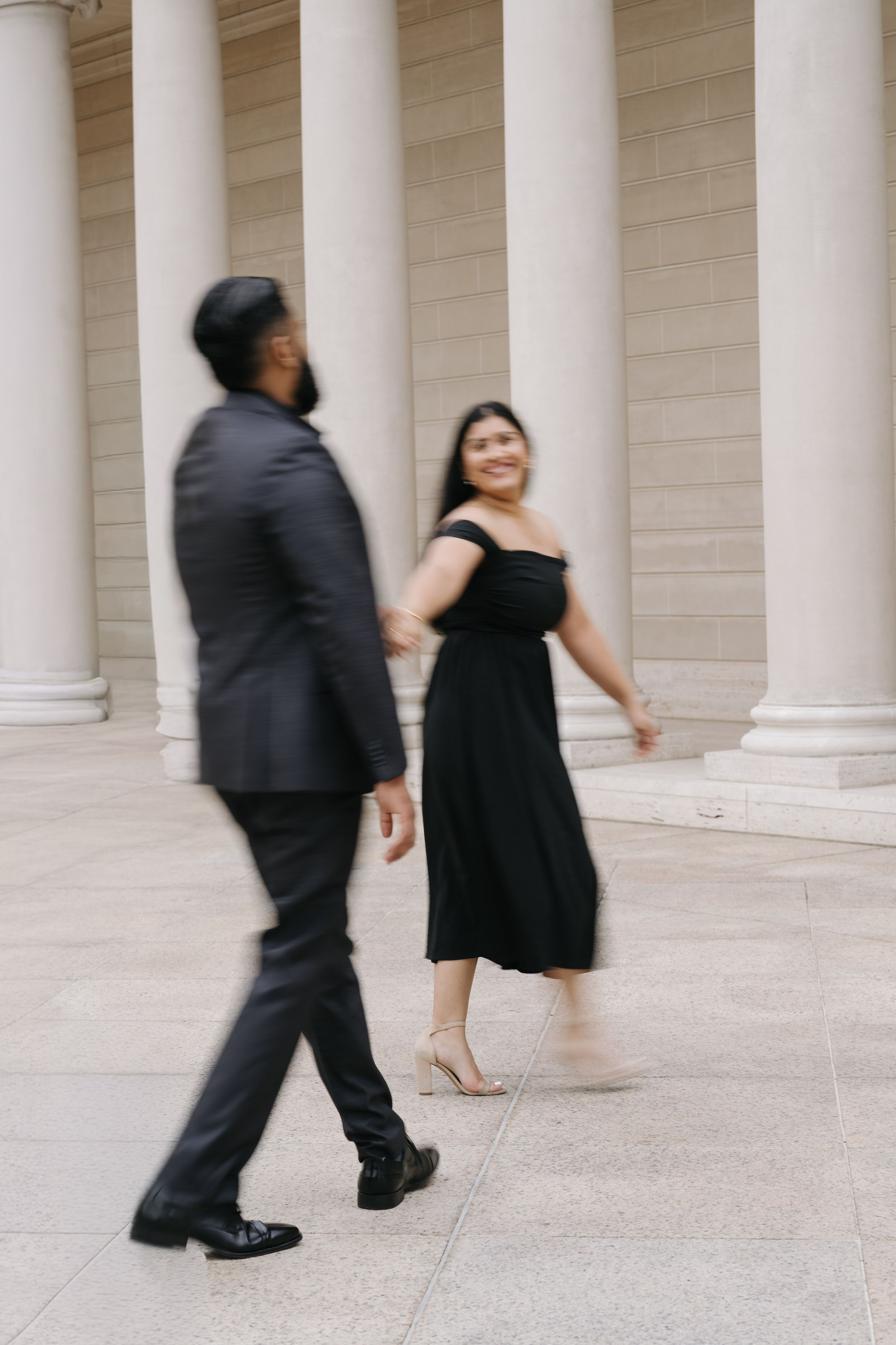 Couple walking hand-in-hand past tall columns, woman in black off-shoulder dress, man in dark suit.