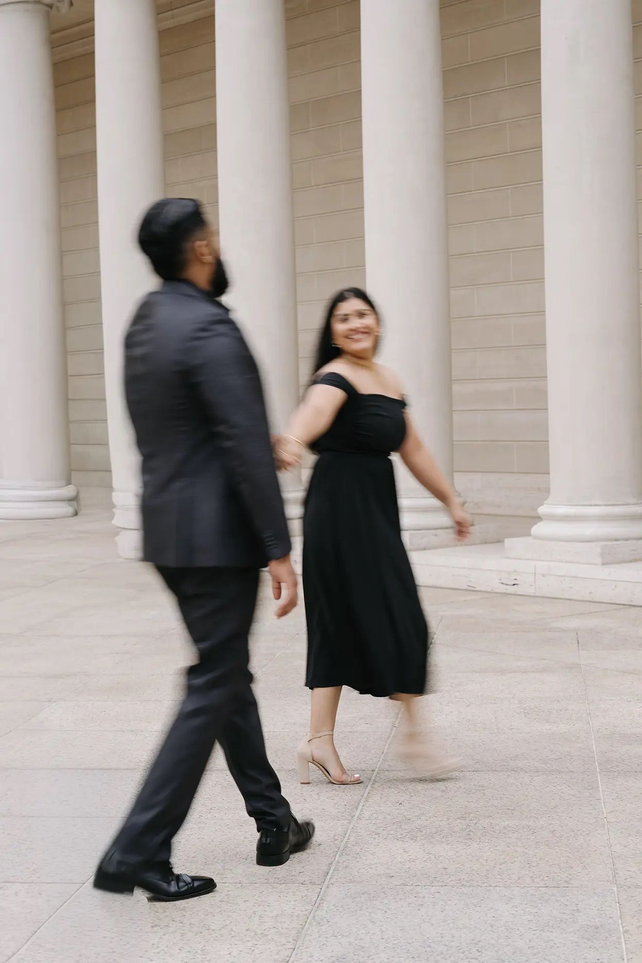 Couple in black formal attire walking among grand columns, motion blur capturing romantic movement.