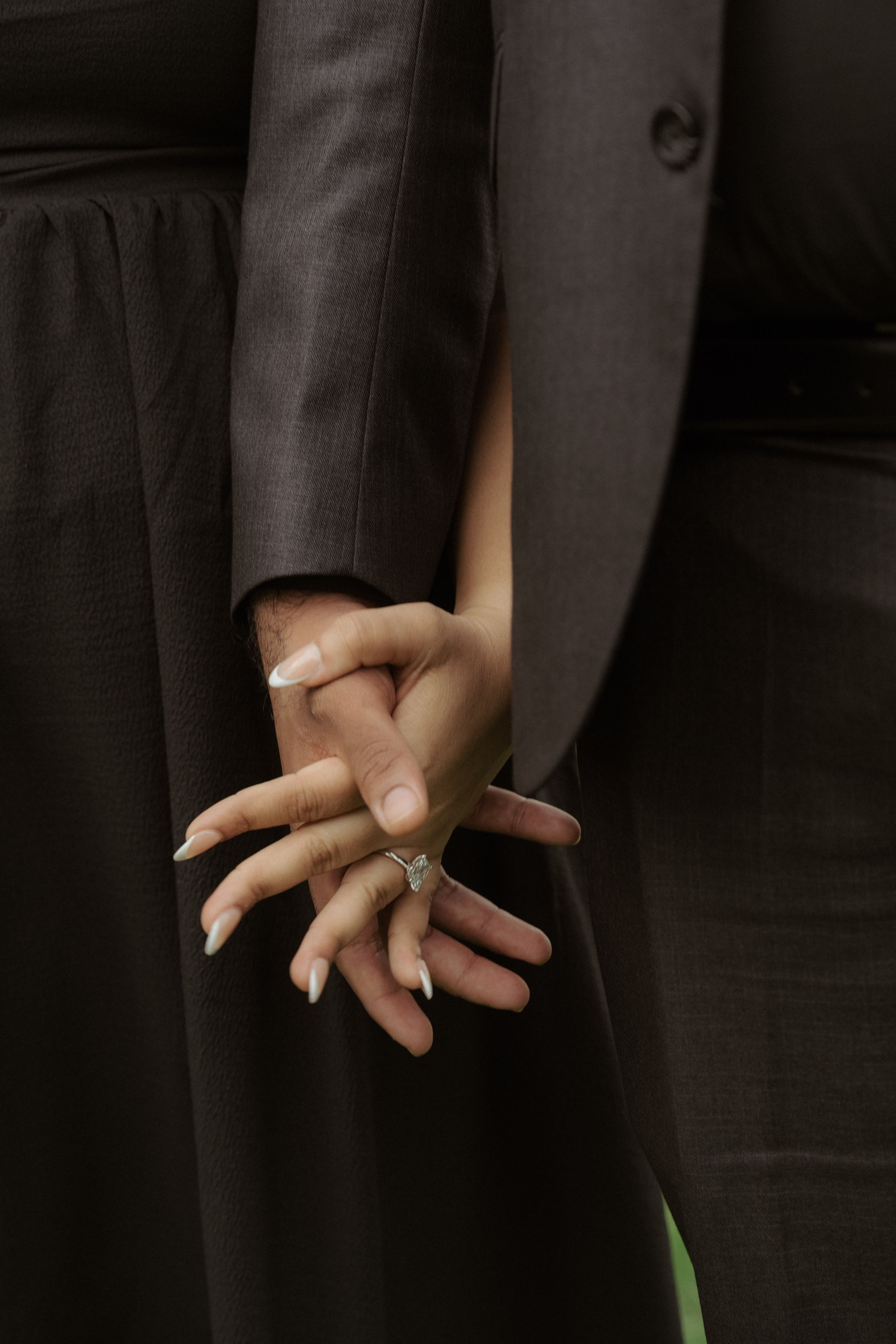 Close-up of engaged couple holding hands showing diamond engagement ring in matching dark formal attire.