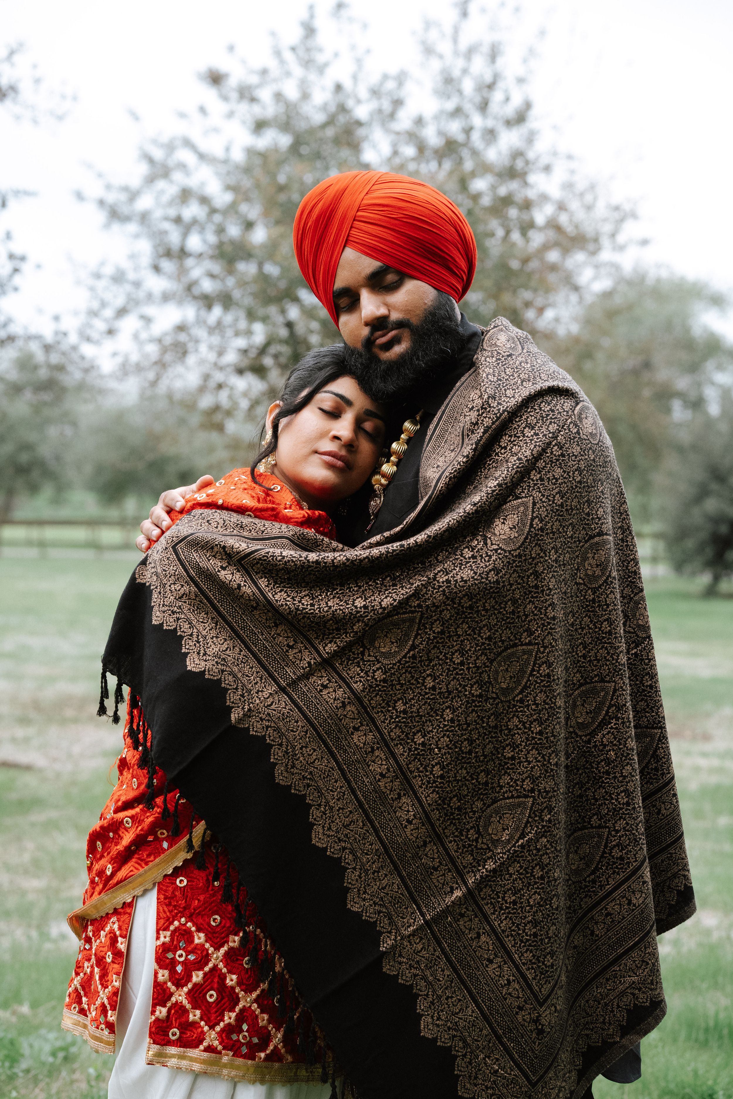 Sikh couple wrapped in ornate shawl outdoors, man wearing red turban embracing woman in red embroidered outfit.