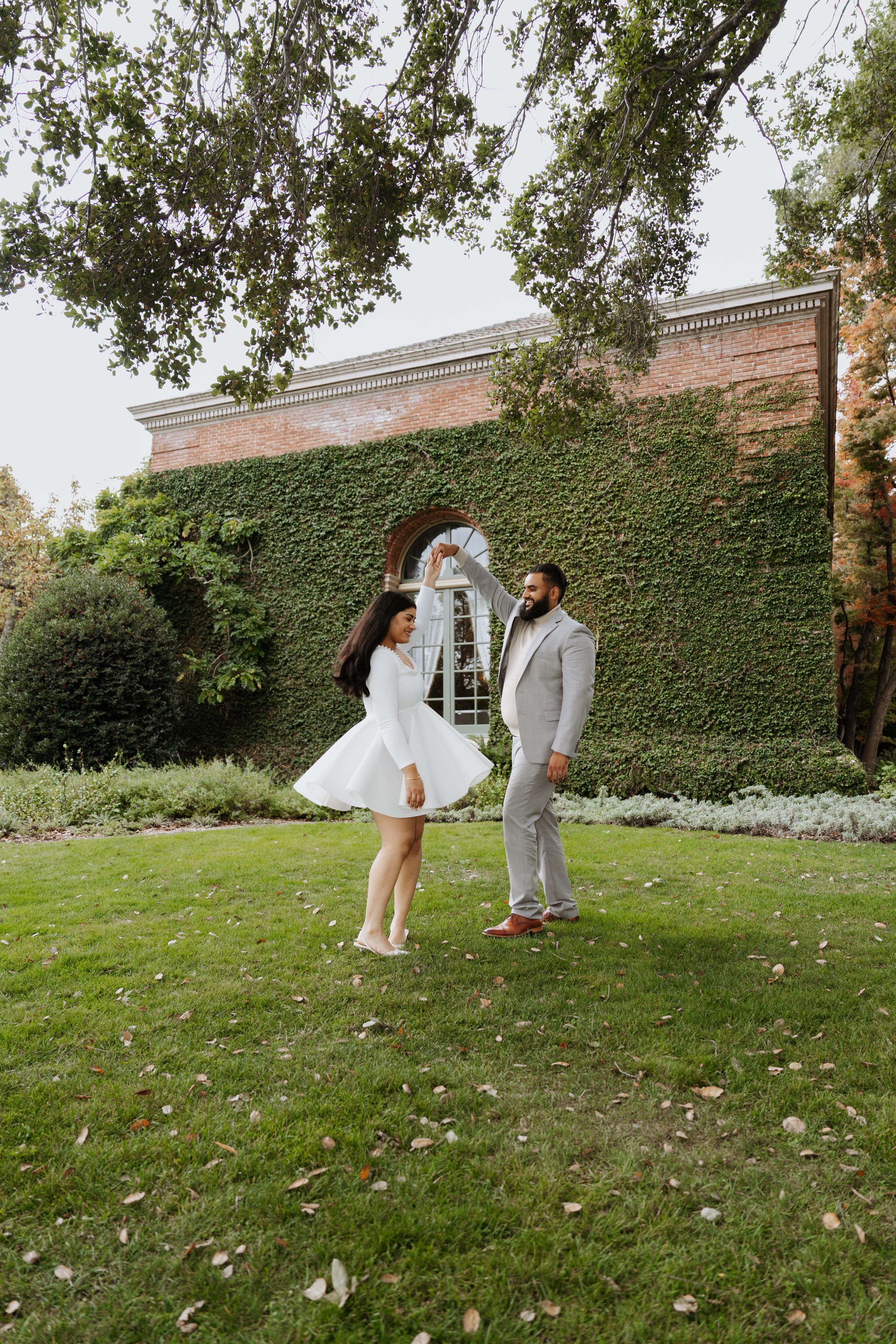 Couple dancing on green lawn in front of ivy-covered brick building during outdoor engagement photo session.