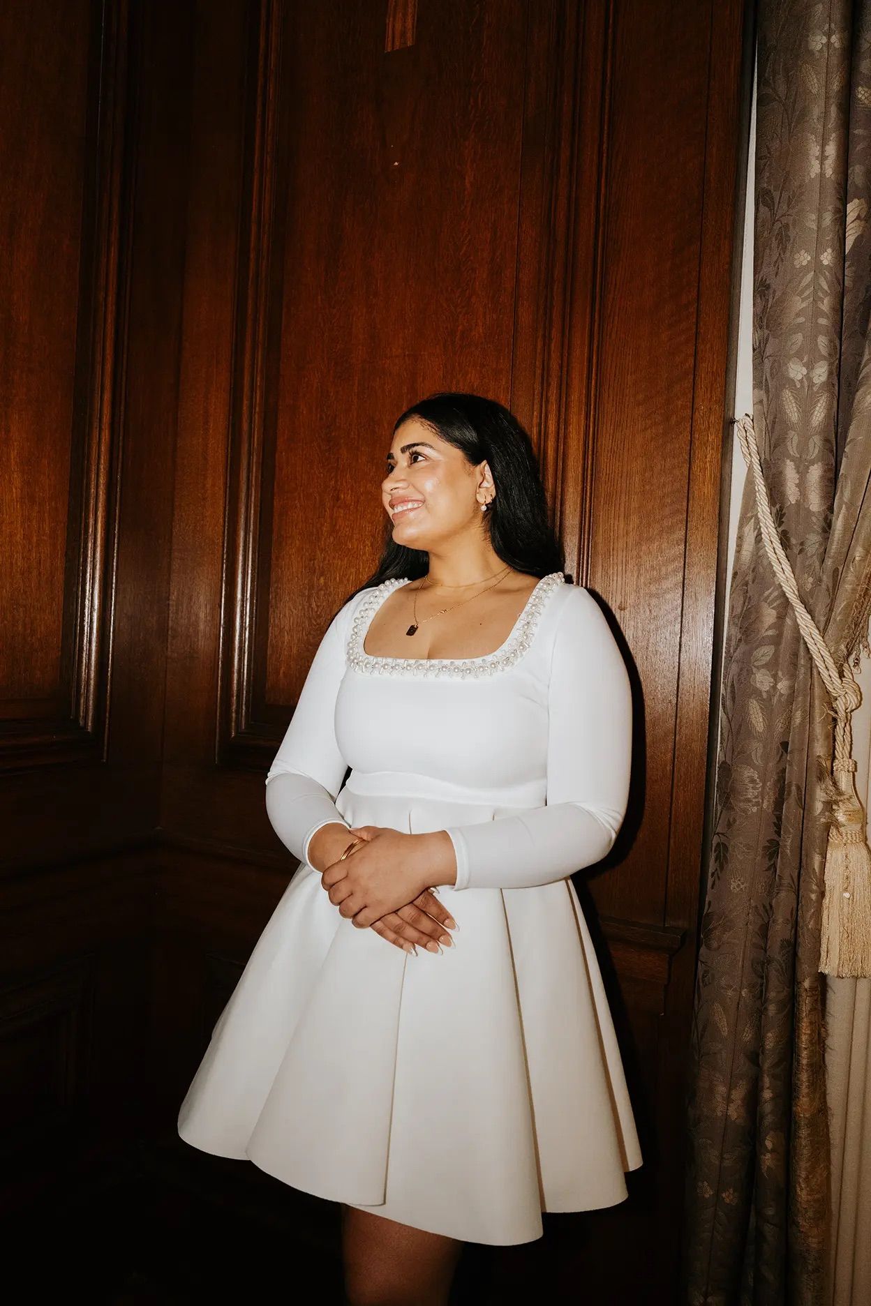Bride in white long-sleeve mini dress with pearl trim smiling in front of wooden paneled wall.