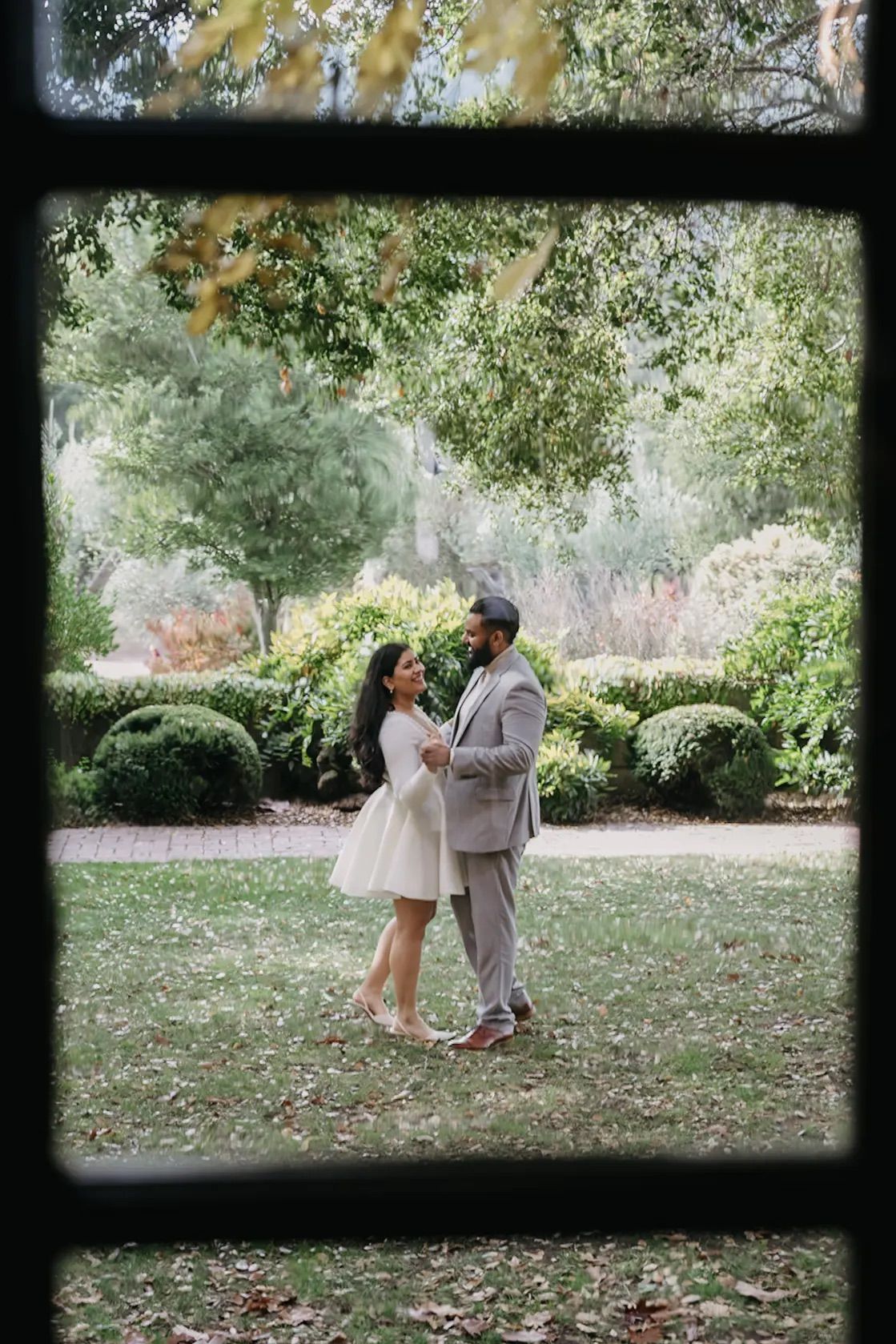 Couple in wedding attire dancing together in lush garden viewed through window frame.