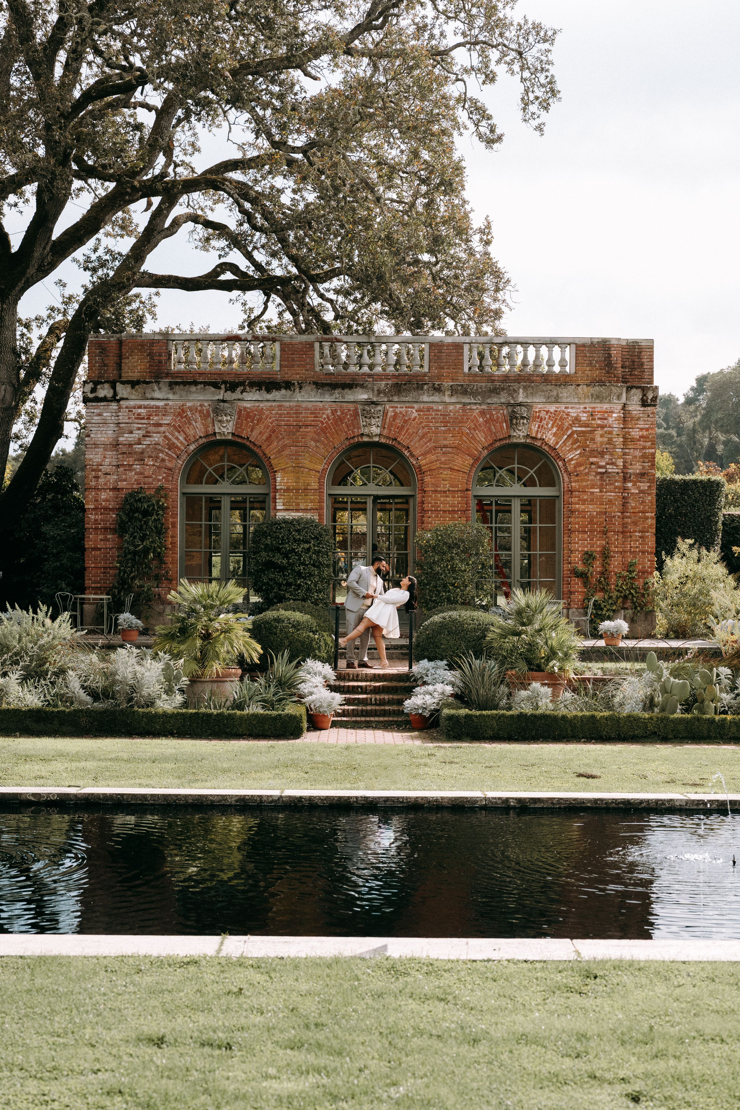 Couple embracing near brick building with arched windows reflected in formal garden reflecting pool.