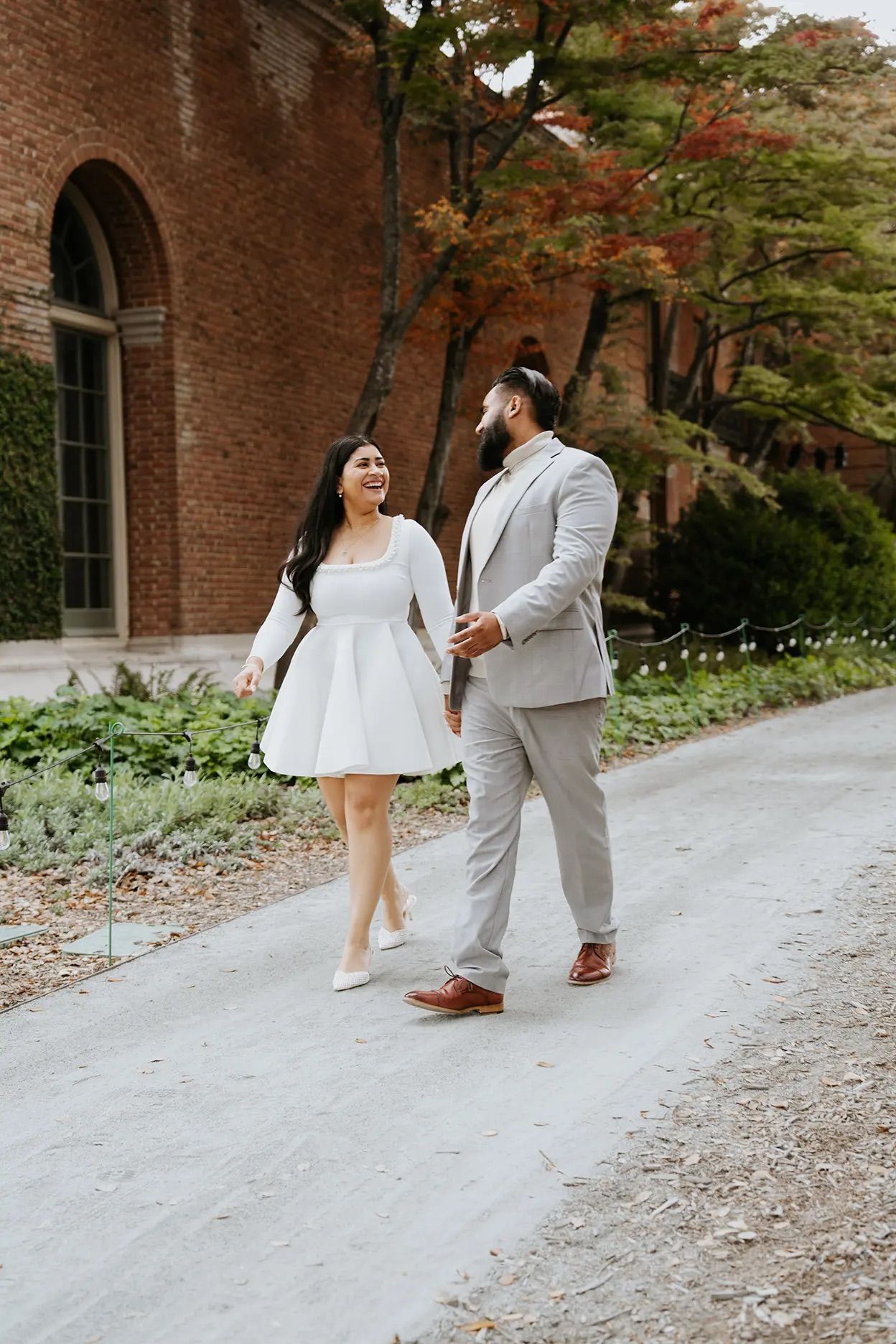 Couple in white dress and grey suit laughing while walking hand-in-hand at outdoor engagement photoshoot.