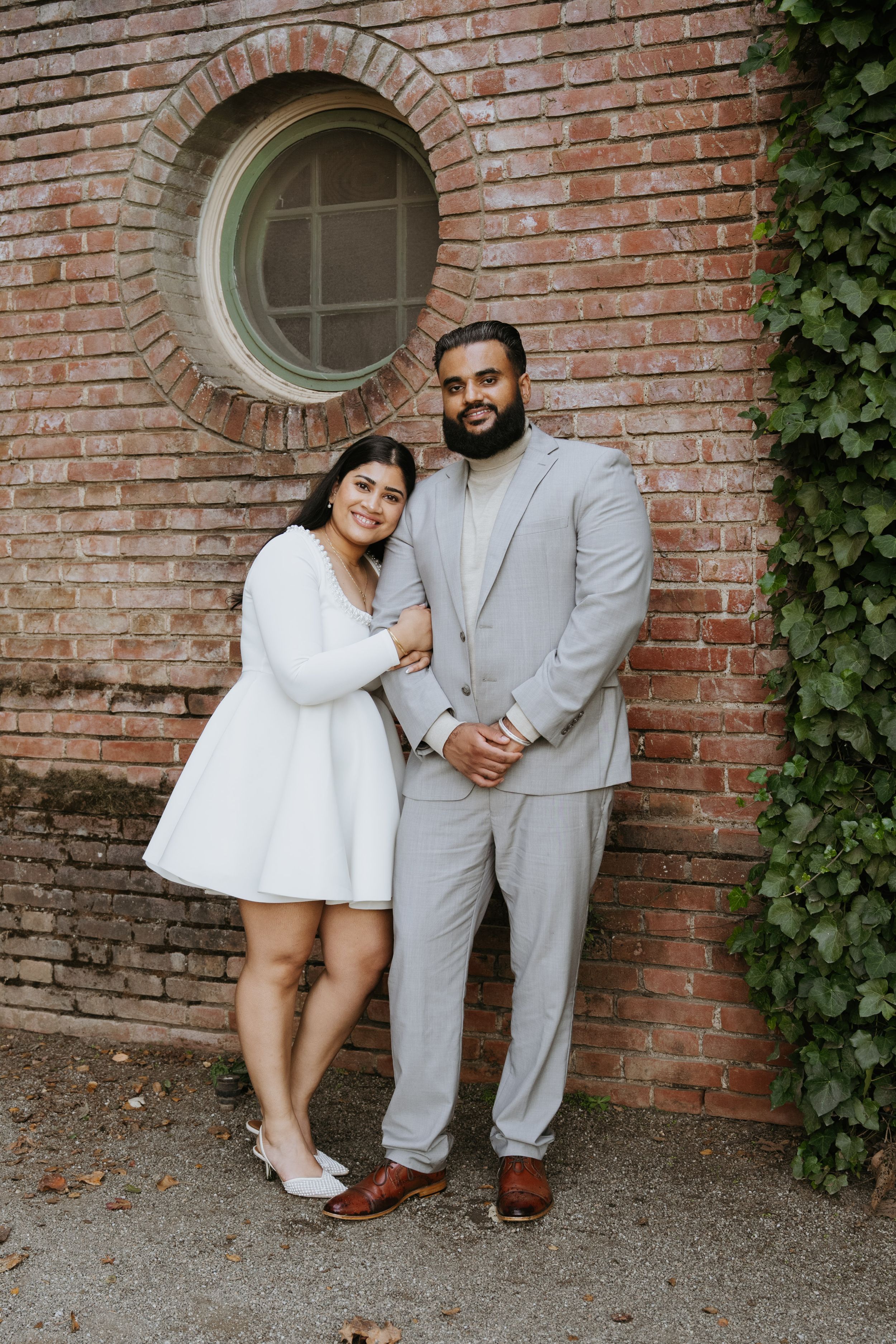 Couple in white dress and gray suit posing against brick wall with round window and ivy for engagement photo.