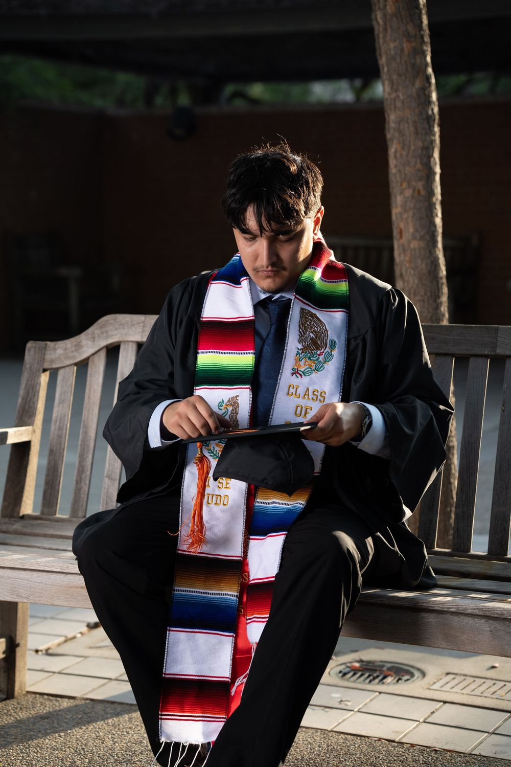 A graduate wearing a colorful Mexican serape stole and graduation gown sits reading on a wooden bench.