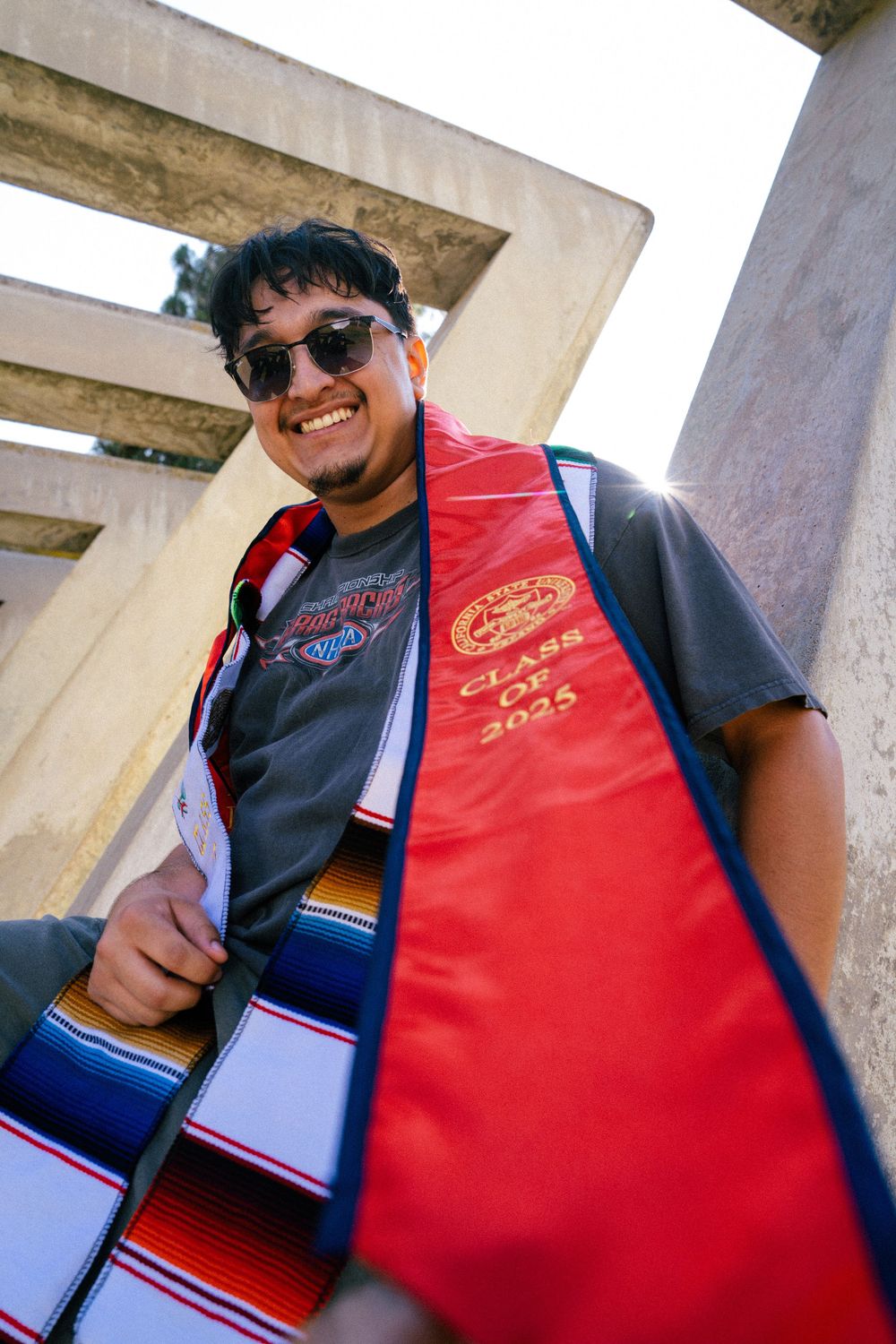 A person wearing sunglasses and a red vest poses under concrete architecture, decorated with a colorful serape-style scarf.