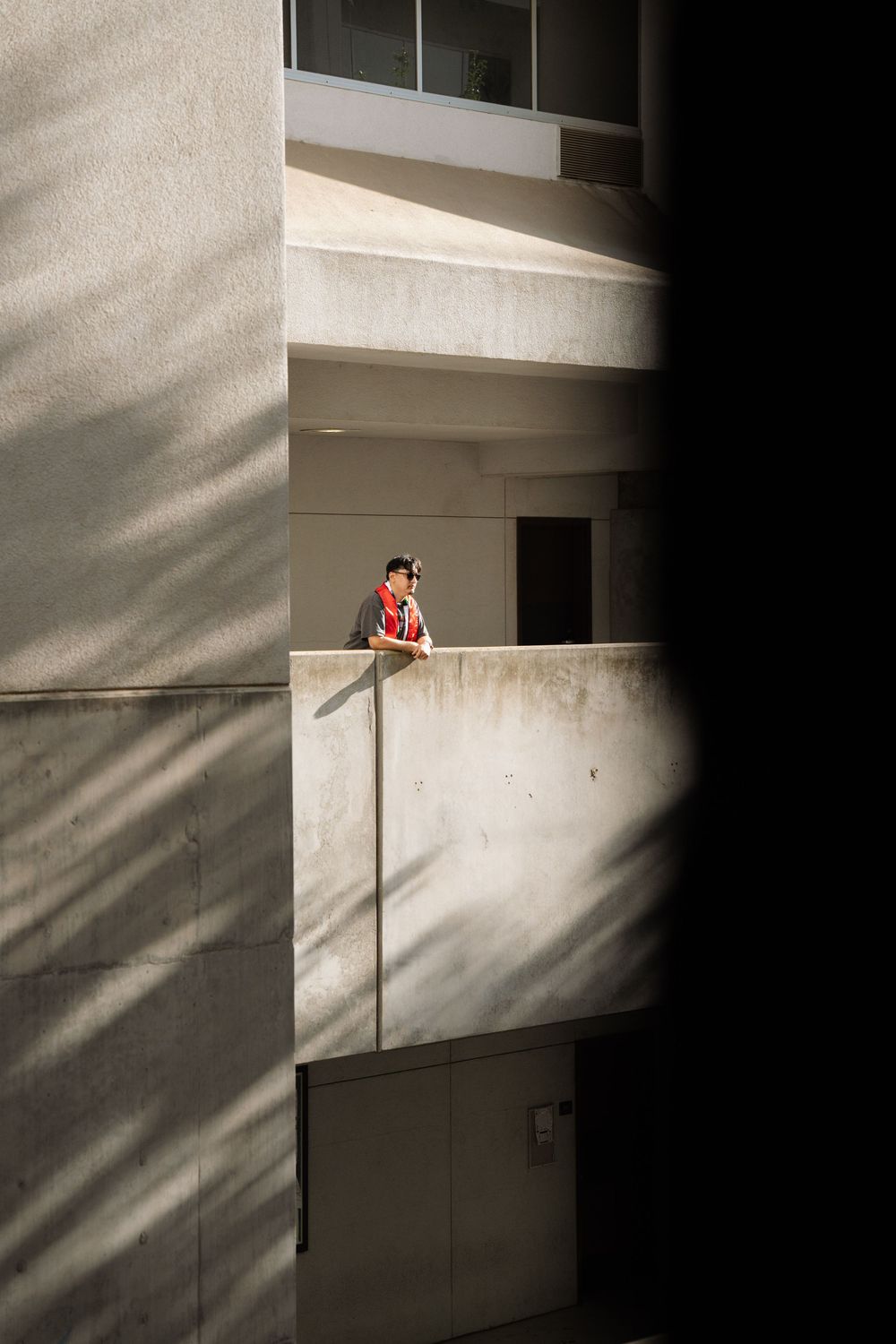 Person in red shirt leans on concrete balcony railing in modern building with dramatic light and shadow contrast.