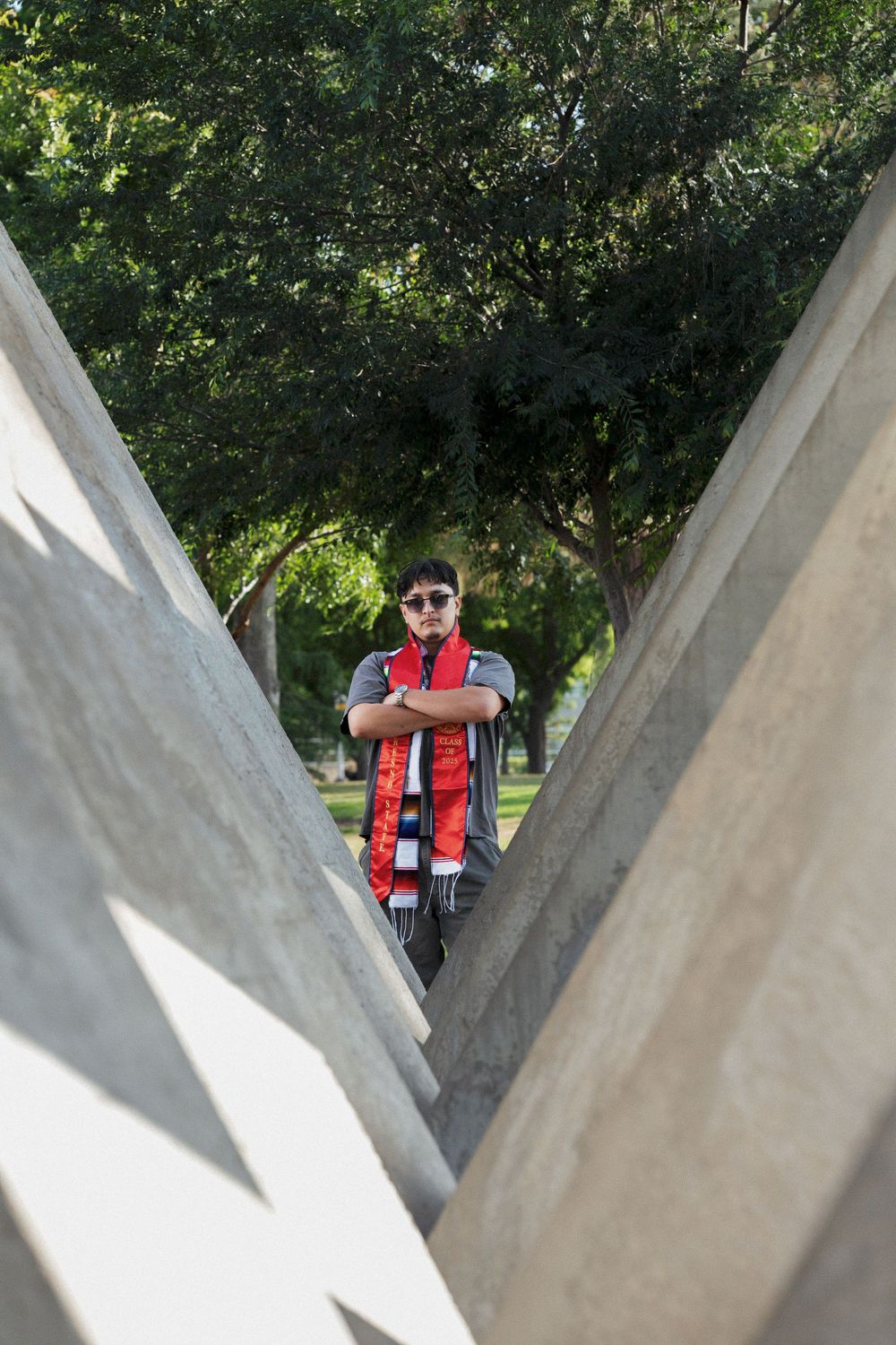 A person in red and black attire stands framed between two concrete walls outdoors with trees in the background.