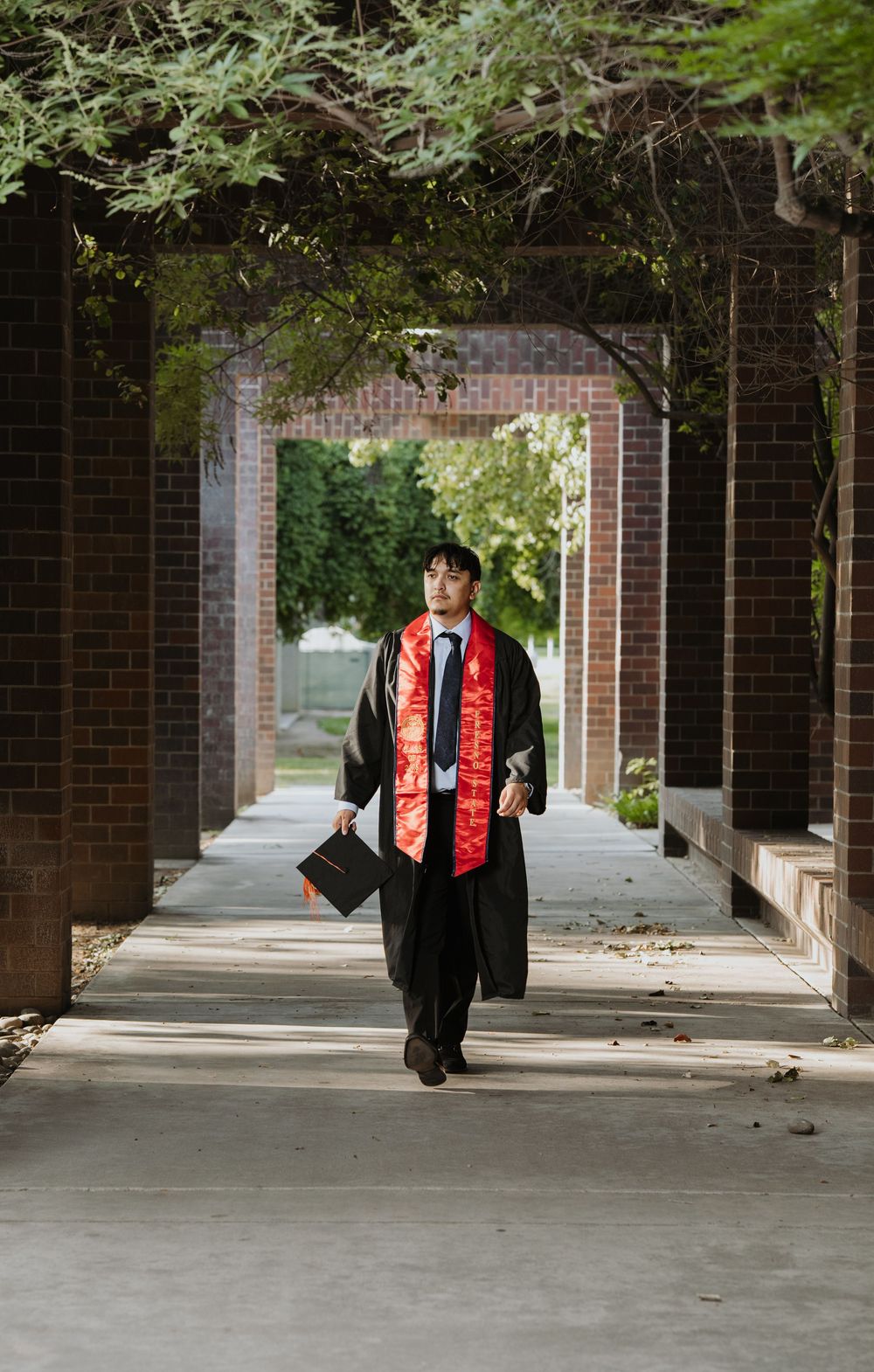 Graduate in cap and gown walking through brick archway corridor holding diploma at university graduation ceremony.