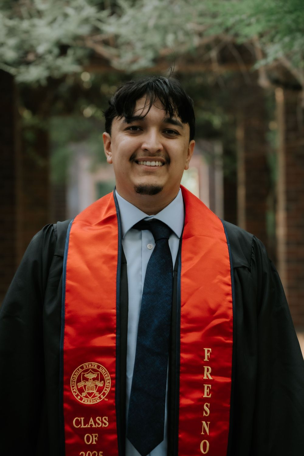 Graduate wearing black graduation gown with red stole and sash stands outdoors during commencement ceremony.