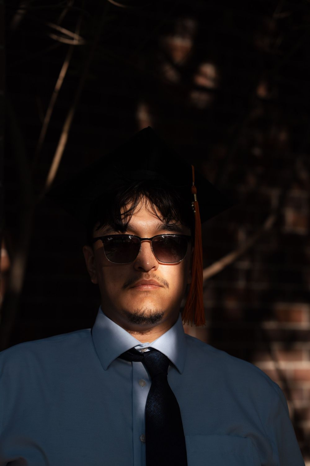 Stylish figure in blue suit and black tie wearing sunglasses stands in dramatic shadow-lit setting.