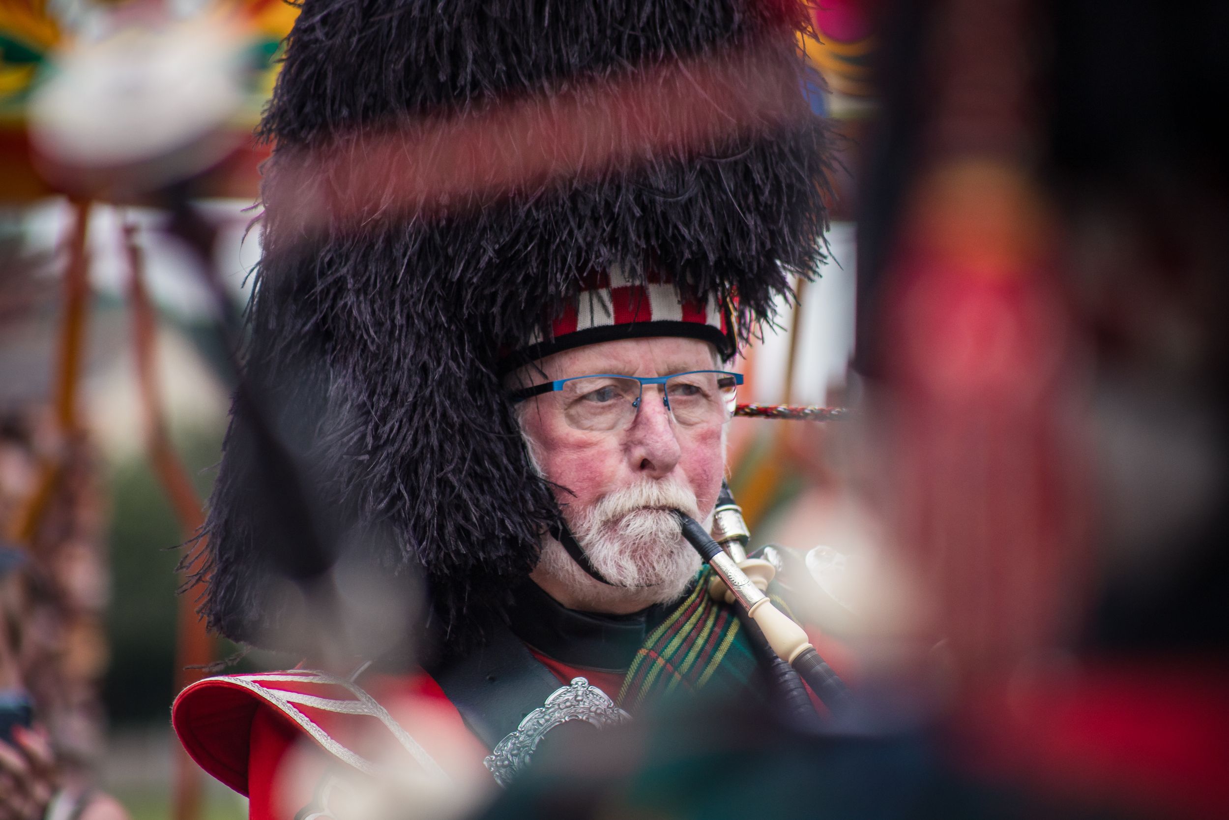 Photo of a man with a white beard bagpiping, shot through the pipes of another piper to create an artistic looking viewpoint