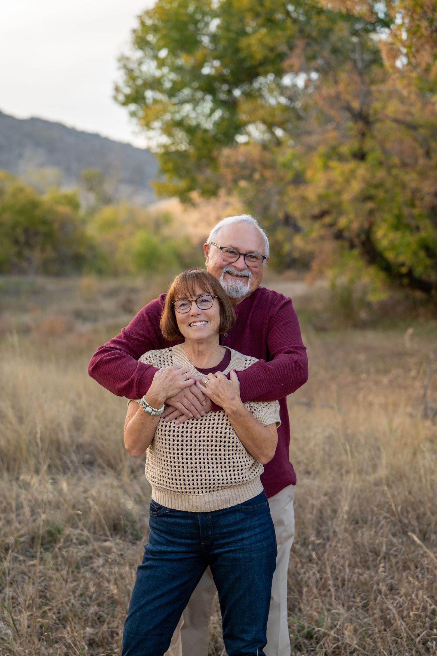Joyful couple embraces in autumn field with mountains and fall foliage in background.