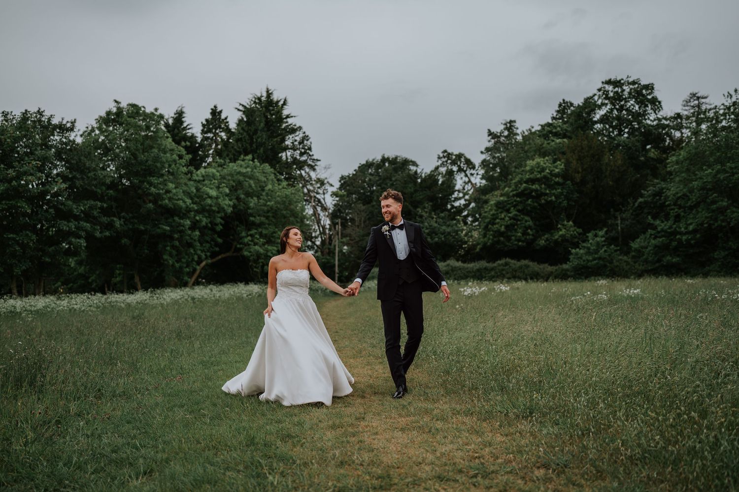 Wedding couple stands in a pastoral field at sunset with a friendly white goat beside them. Woodlands Hothorpe