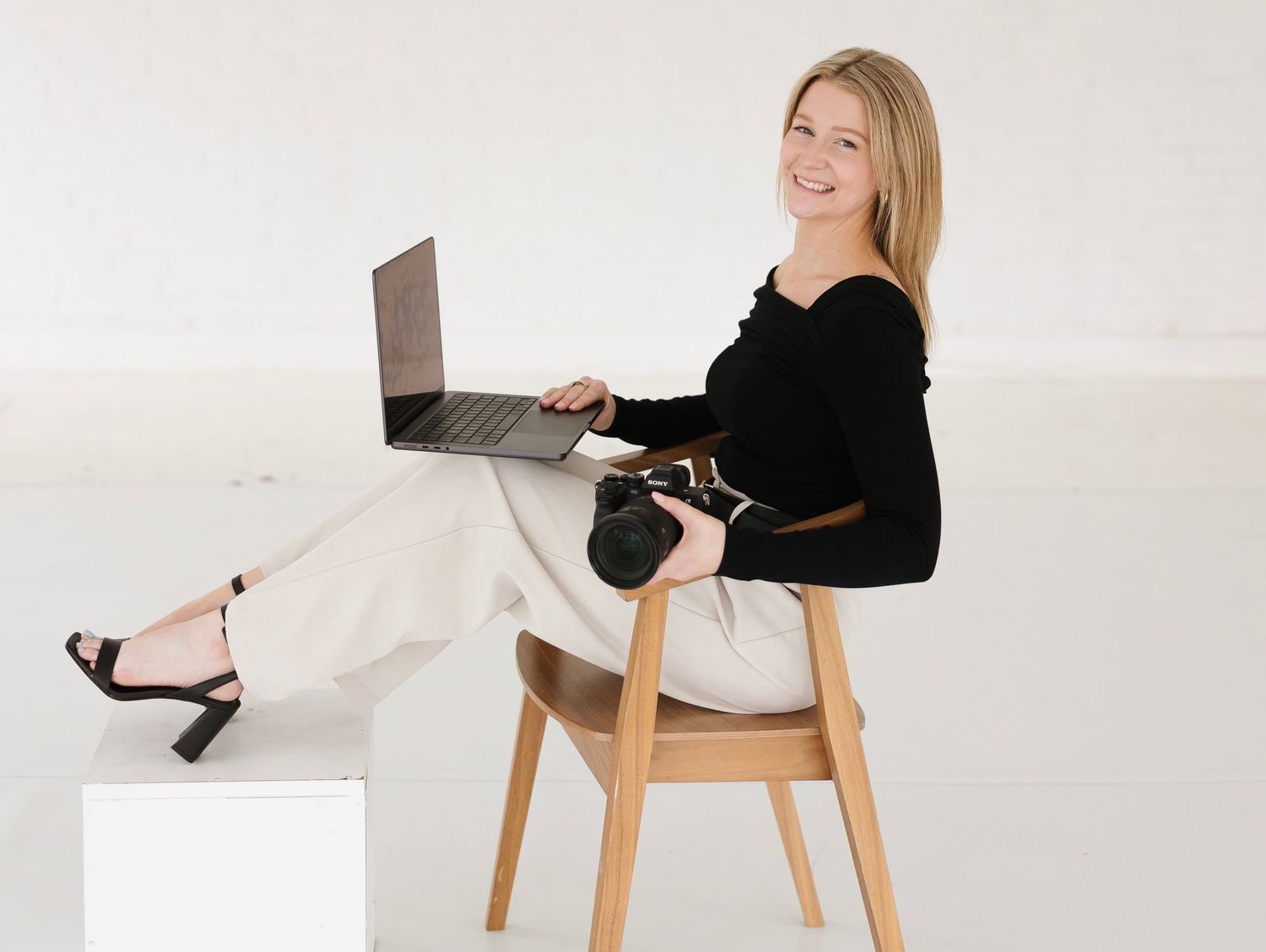 A person in a black sweater and white pants sits comfortably on a wooden chair while working on a laptop.
