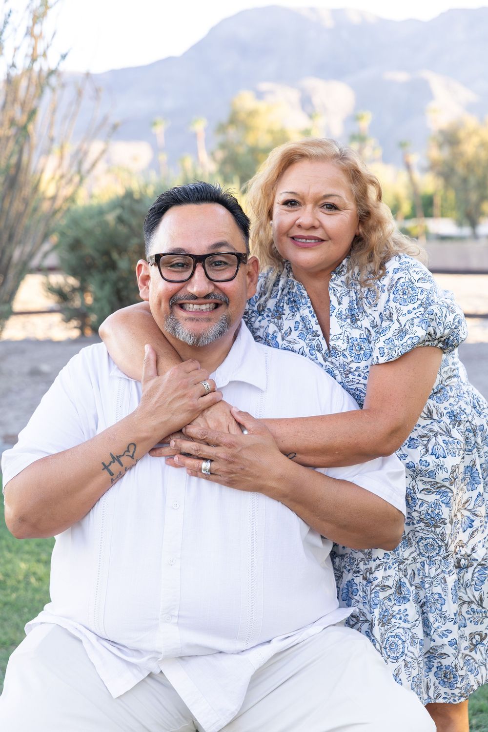 A smiling middle-aged couple poses outdoors, woman in blue floral dress embracing man in white shirt with glasses.