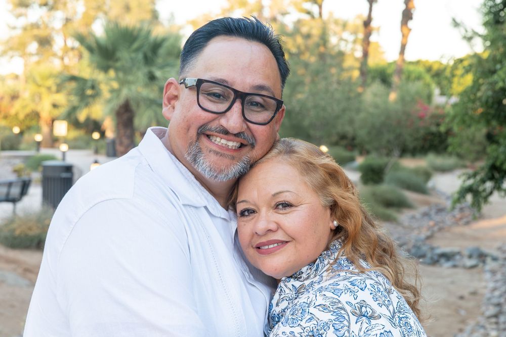A smiling couple embraces outdoors, with the woman hugging the man from behind in a lush garden setting.