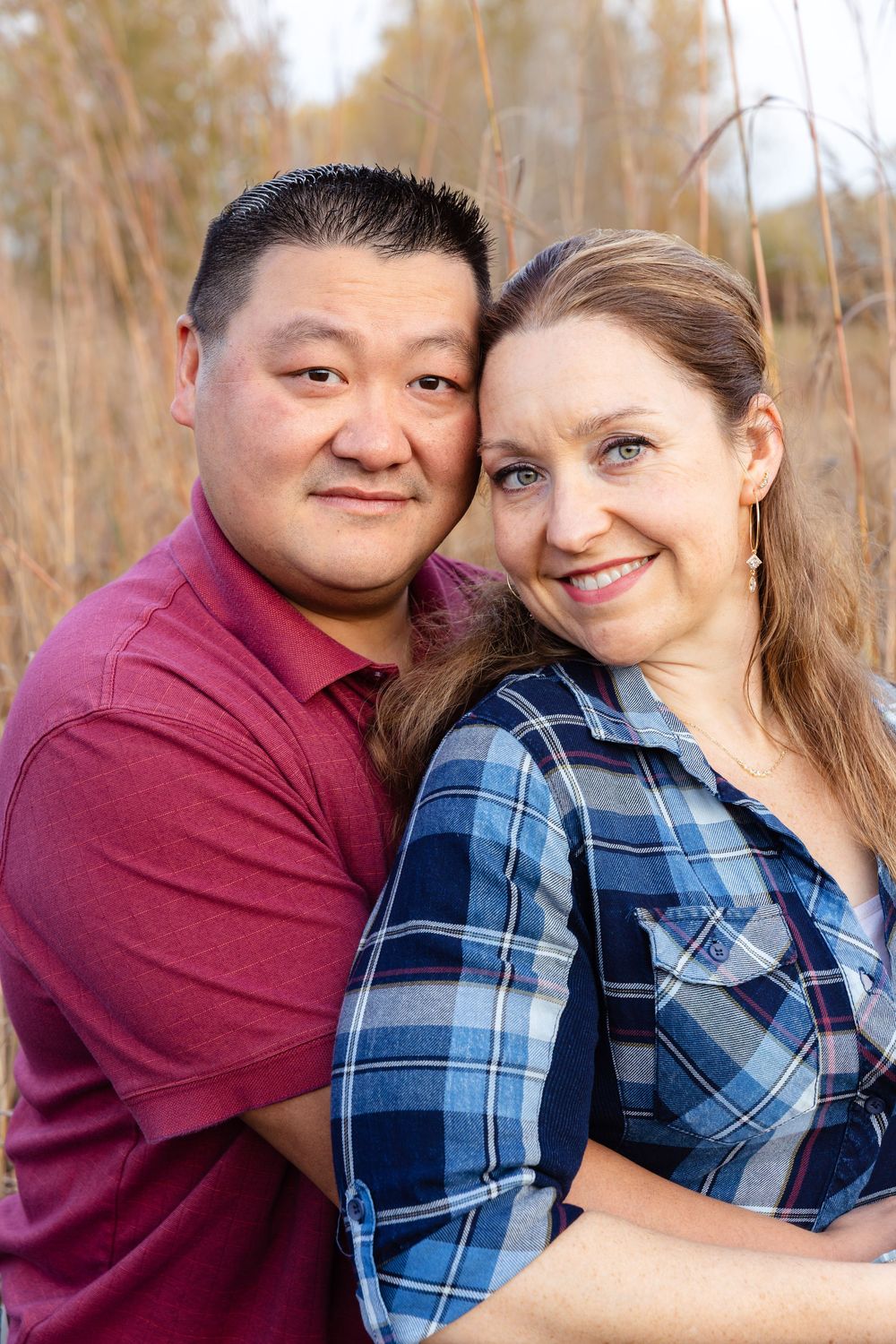 A happy couple poses together outdoors in casual attire with autumn foliage in the background.