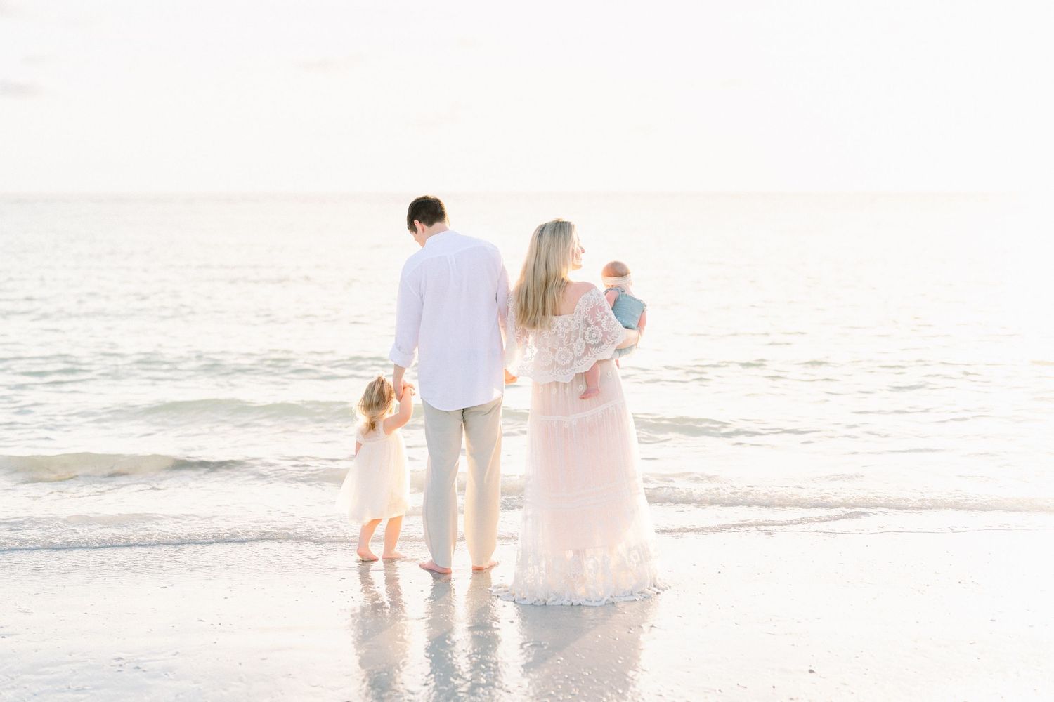 A black and white photo of a family holding hands walking down the beach with rocks in the background with a sea wall at golden hour.