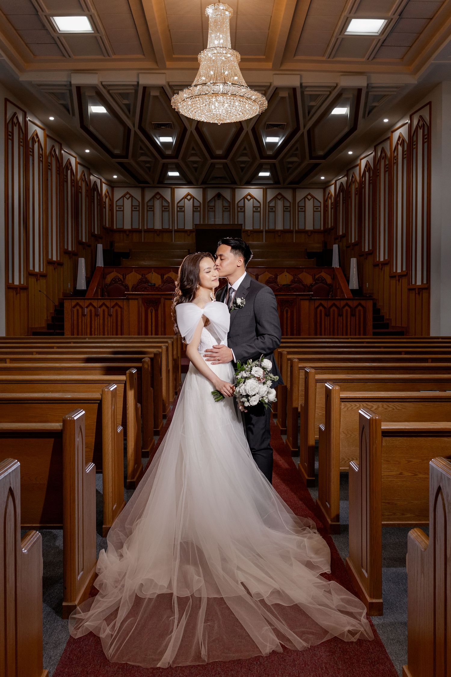 Elegant couple in all-white attire poses between classical columns with ornate lace train.