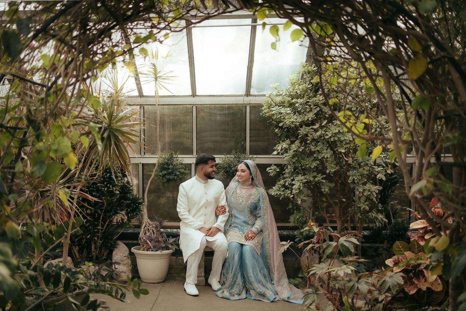 A couple poses in traditional attire inside a lush greenhouse filled with tropical plants.