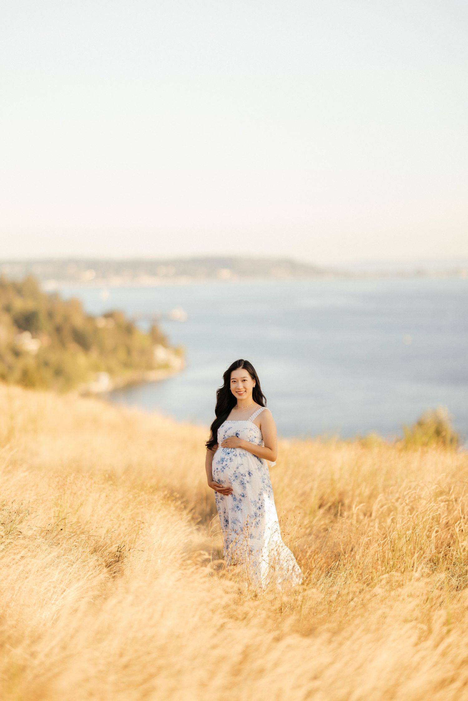 Maternity photo of woman in floral dress standing in golden field with water and coastline in background.
