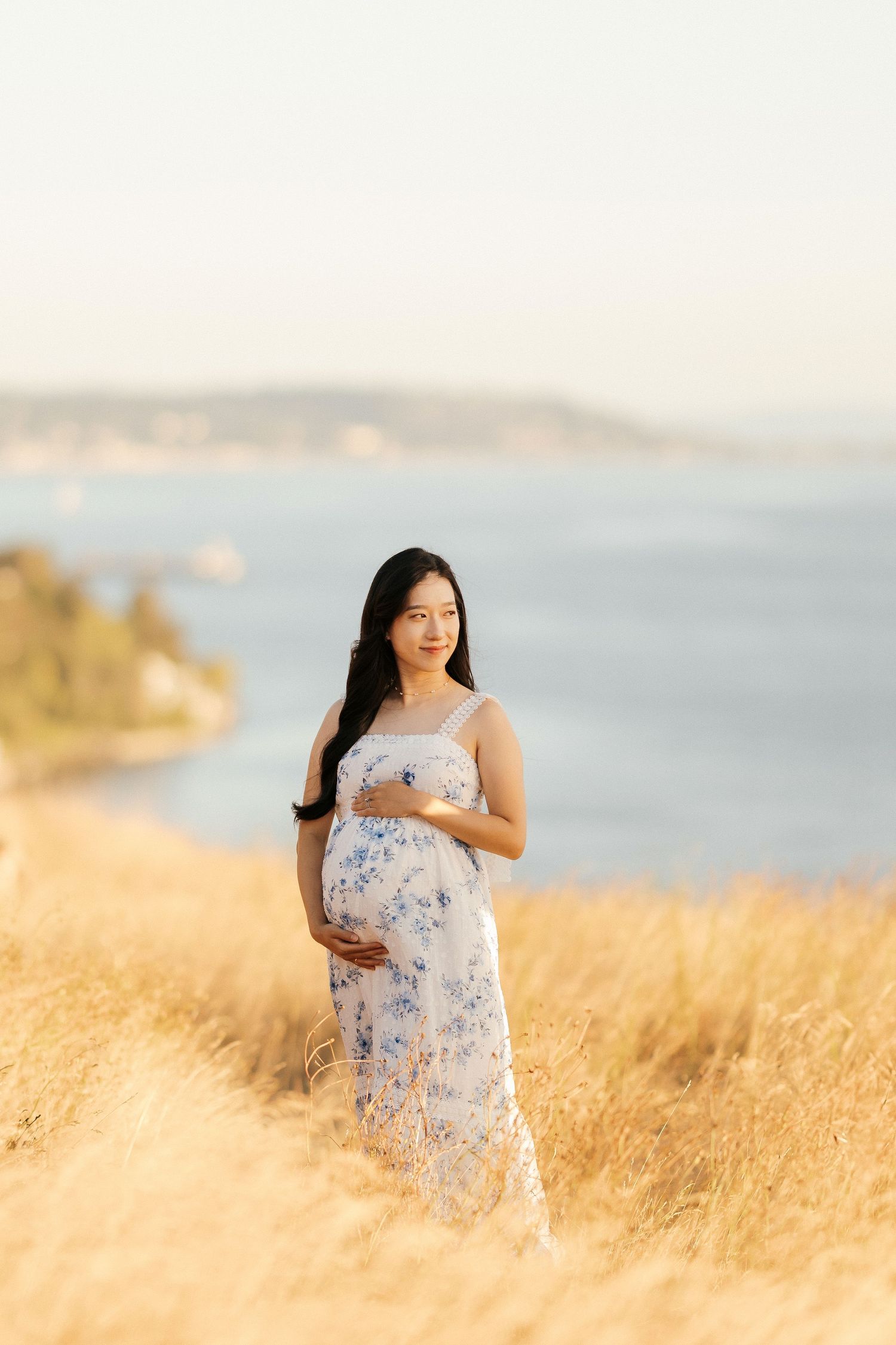 Expectant mother in white floral dress poses in sunlit meadow overlooking coastal waters.