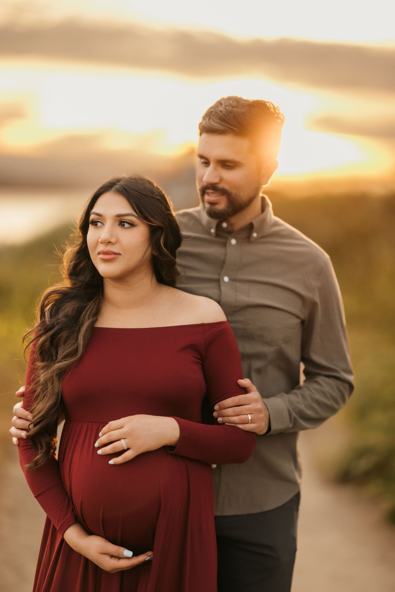 Sunset maternity portrait with couple in burgundy dress and tan shirt against glowing sky.