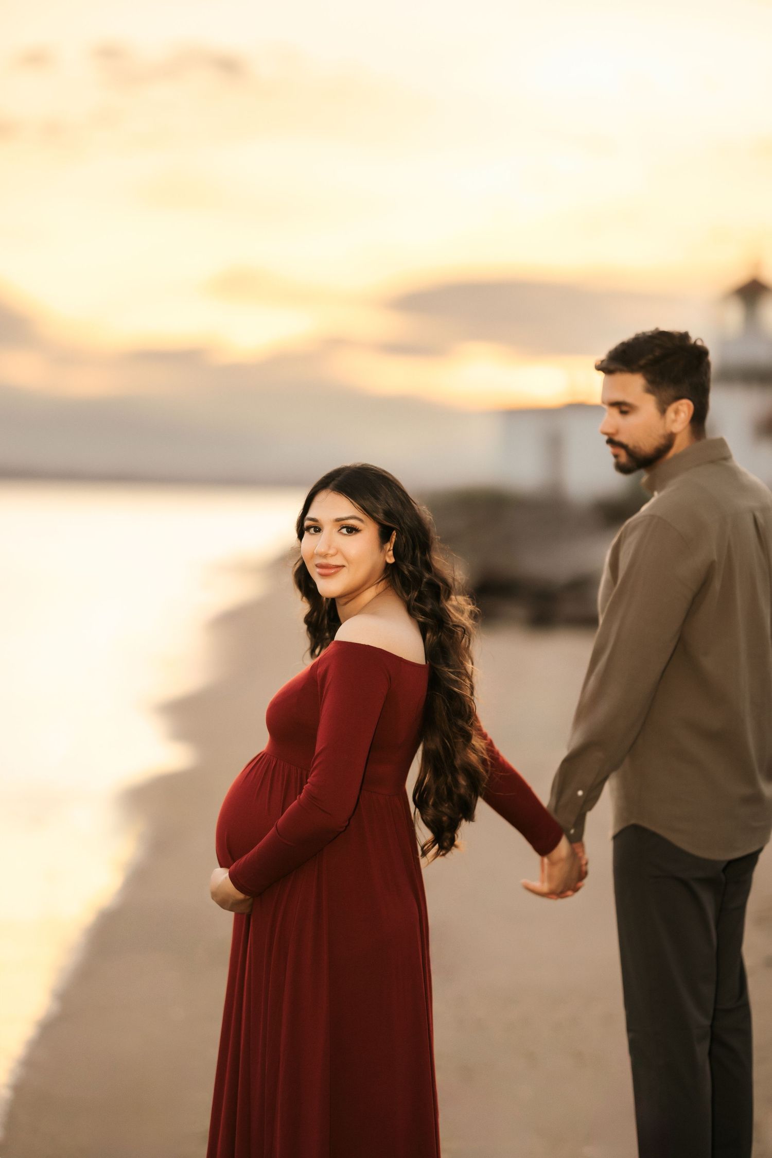 Couple holds hands during sunset maternity shoot with woman in burgundy dress.