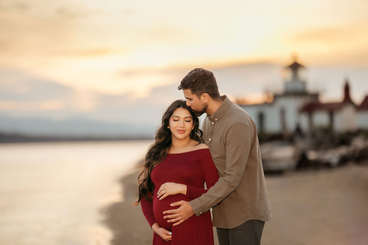 Sunset maternity photo with lighthouse backdrop as couple embraces on beach.
