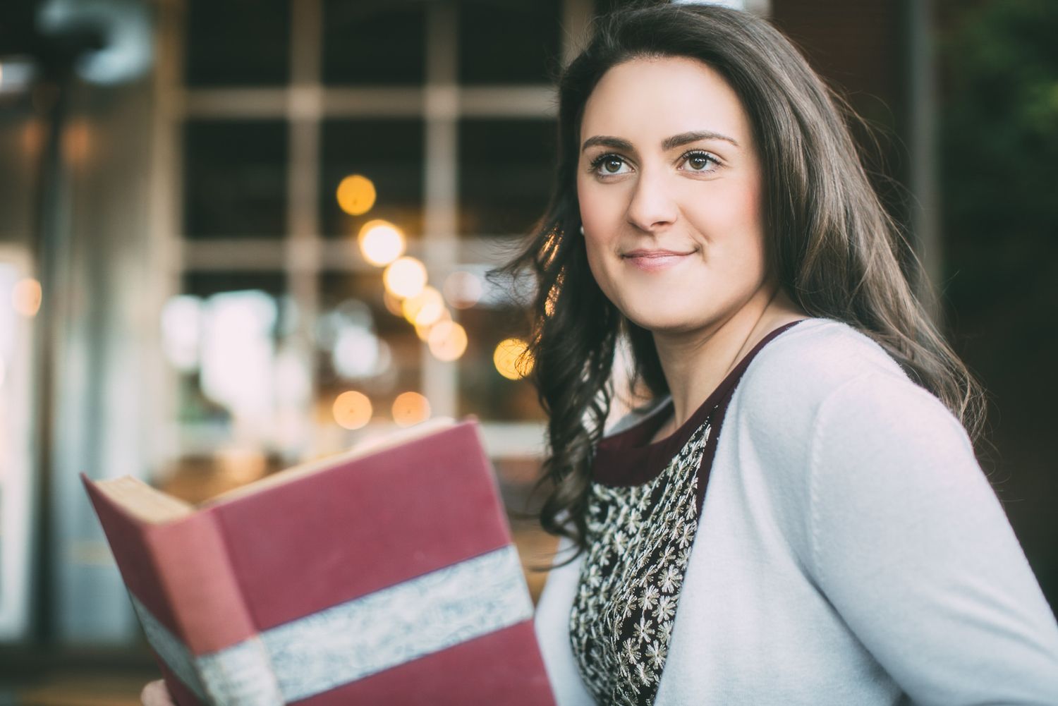 A person in a gray sweater sits at a table smiling near a window with soft bokeh lighting in the background.