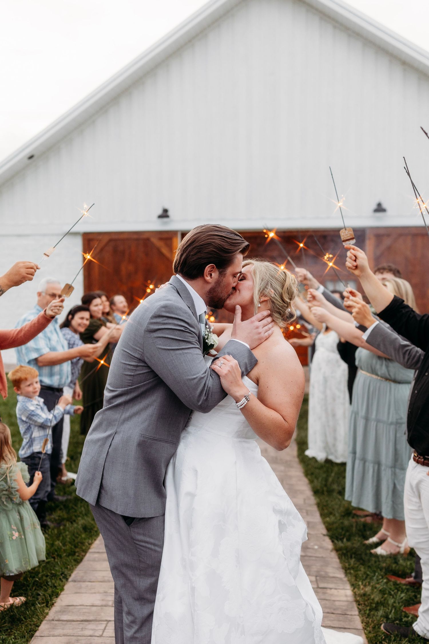 Bride and groom share a kiss during sparkler send-off exit outside white barn wedding venue.