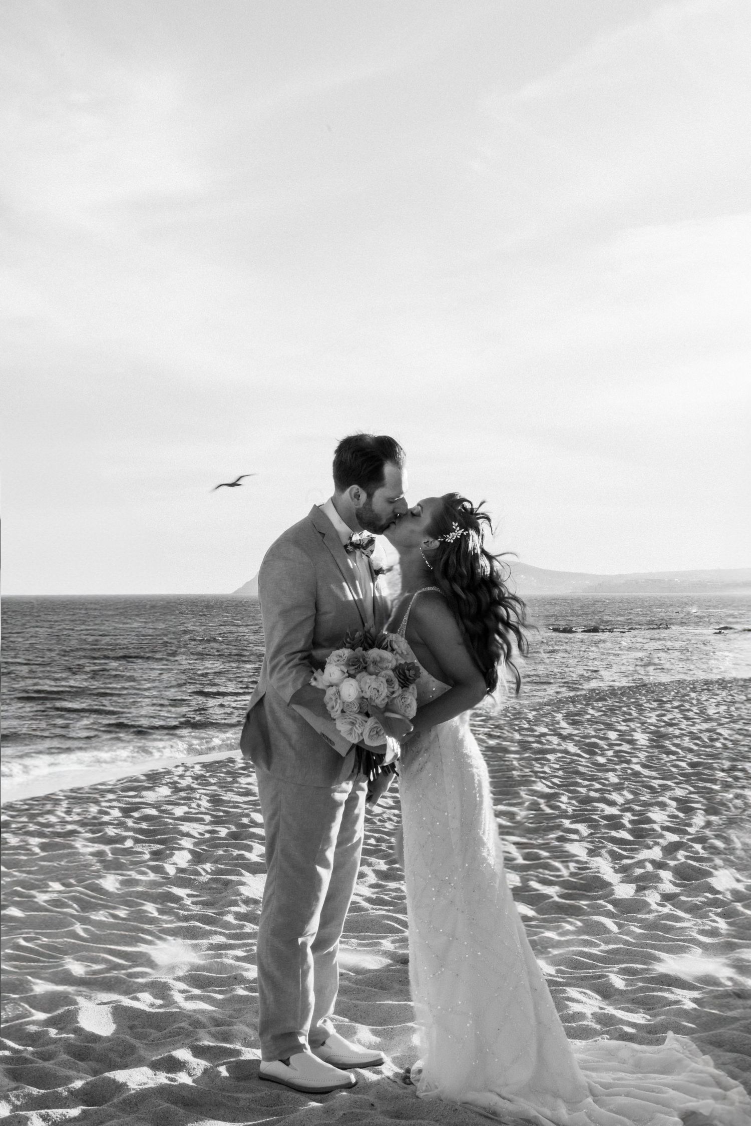 Black and white photo of a bride and groom sharing a kiss on a beach, with the ocean and a bird in the sky in the backgr