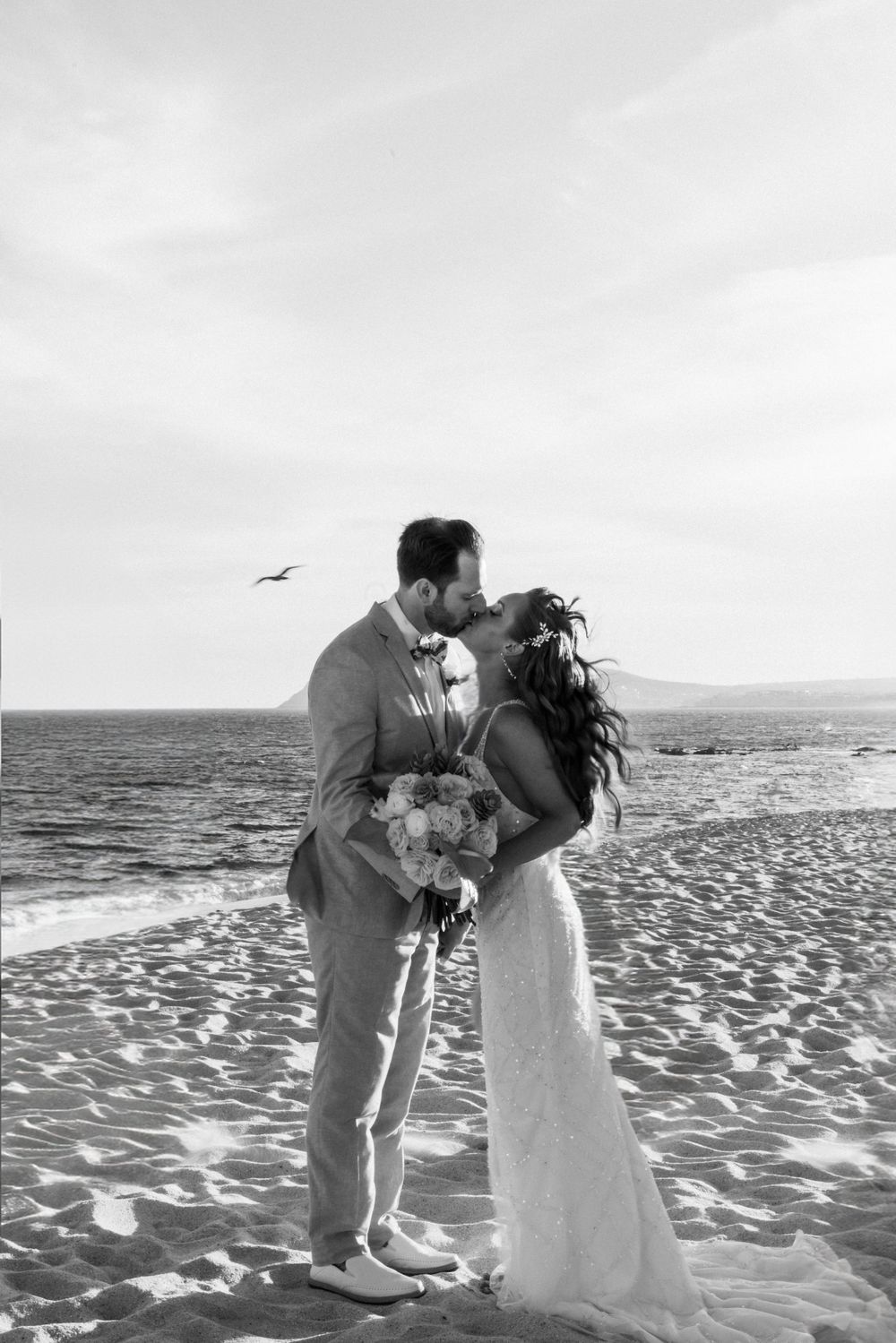 Black and white photo of a bride and groom sharing a kiss on a beach, with the ocean and a bird in the sky in the backgr