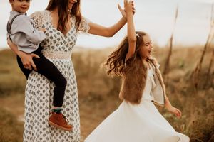 A joyful mother in a floral dress holds a toddler while a laughing girl in white spins outdoors in a golden field.