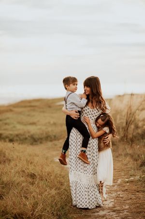 A mother holds her young son while her daughter hugs her on a golden field path, all dressed in boho-style outfits.