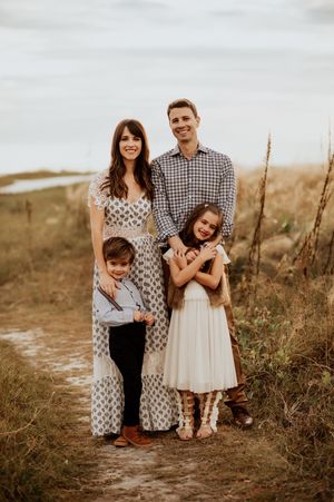 Family of four posing outdoors on a sandy path surrounded by tall grass during golden hour.