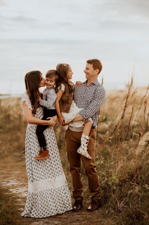 A happy family of four poses outdoors in a field, parents holding two young children, dressed in casual earth-toned outfits.